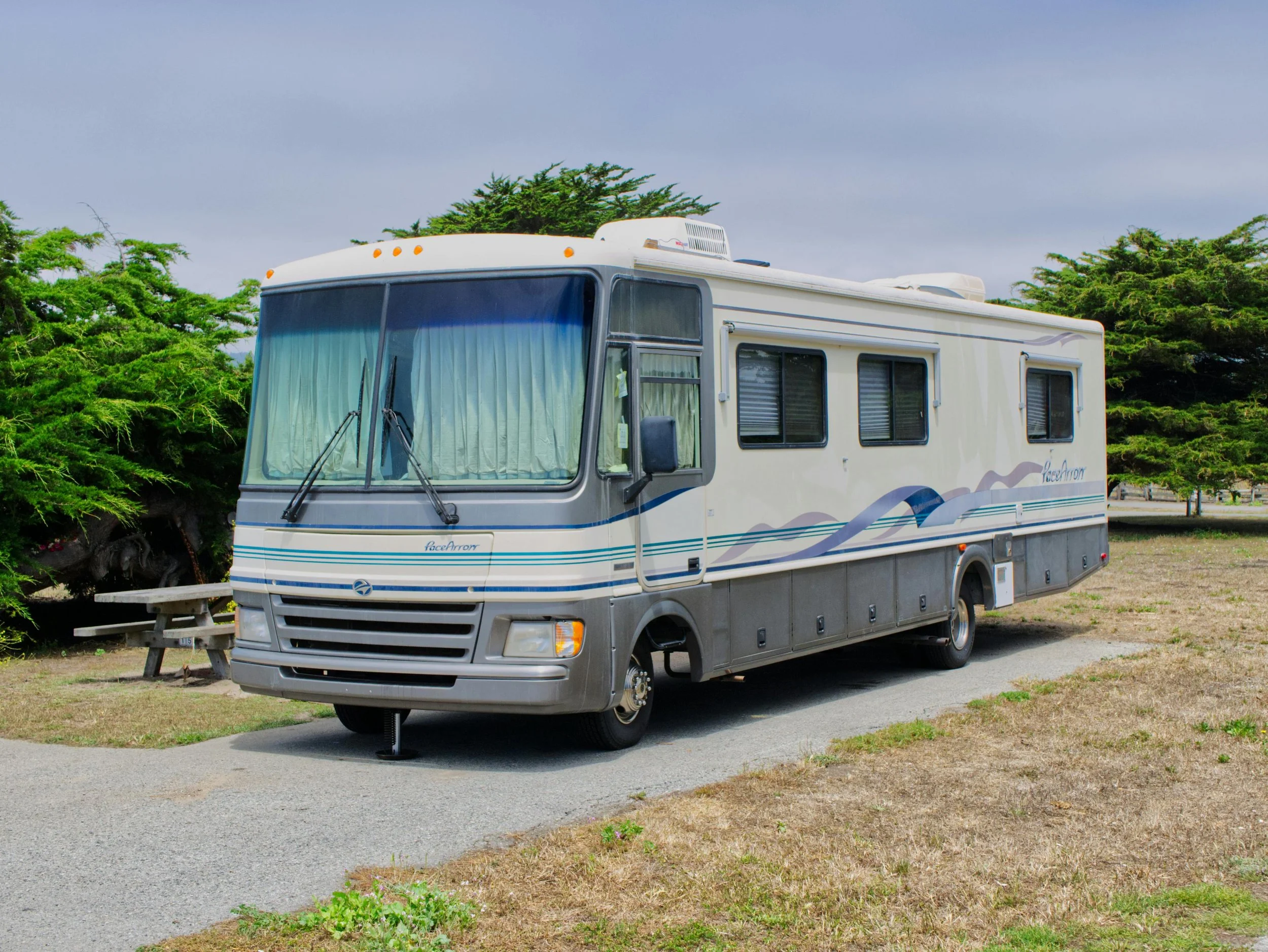 A white motorhome parked on a paved area with trees and grass in the background under a cloudy sky.