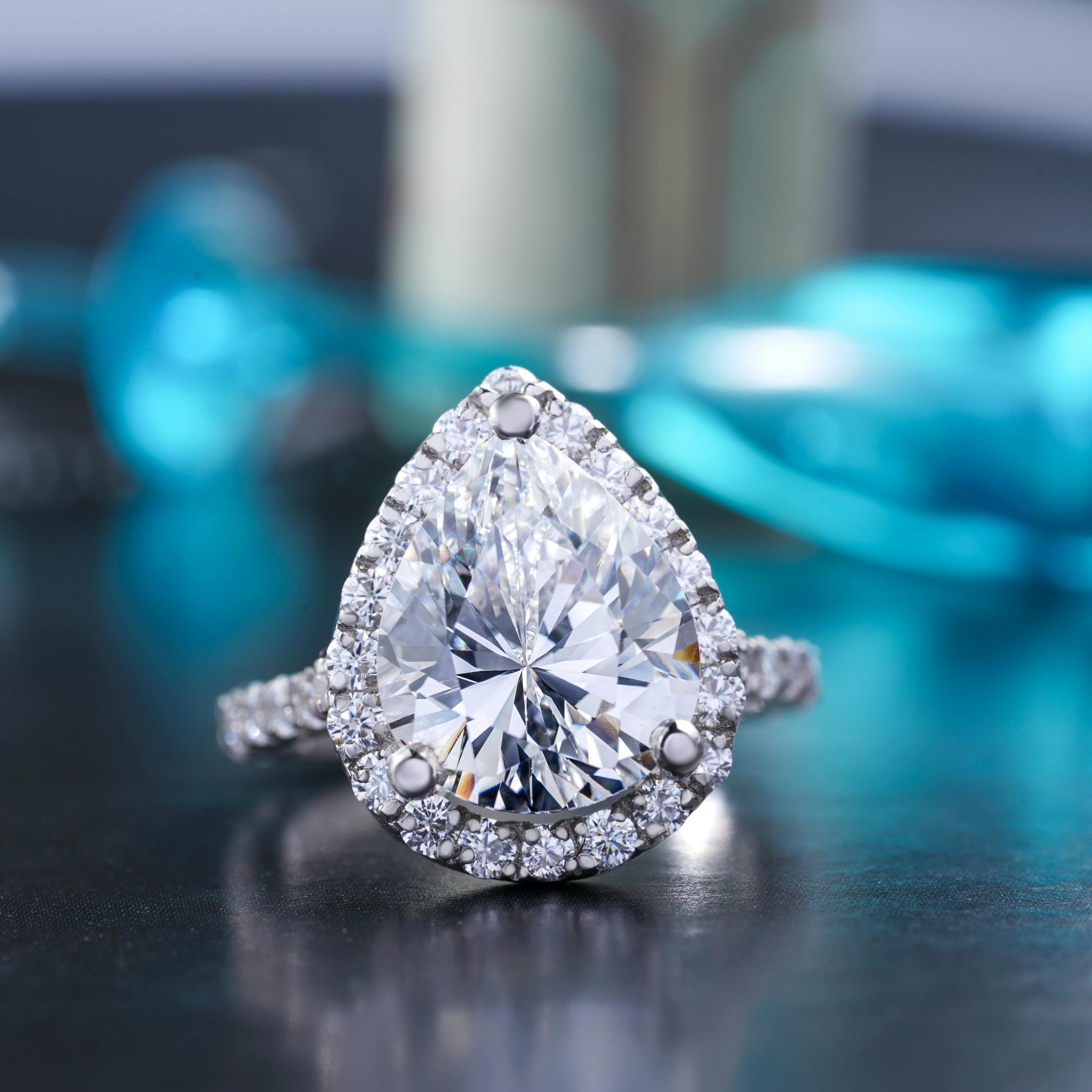 Close-up of a large, sparkling diamond ring on a black surface, with blurred background featuring blue and gray tones.