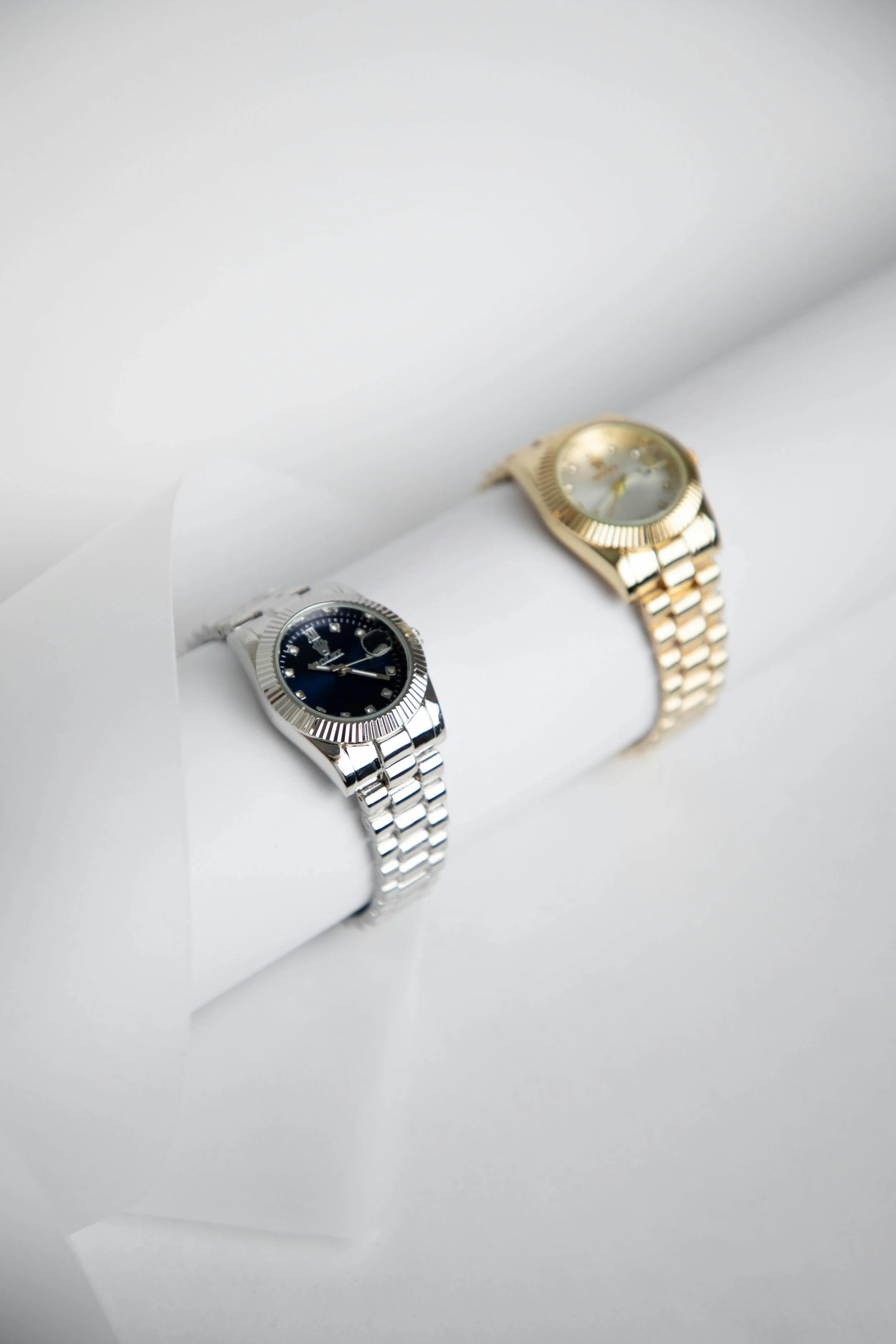 Two wristwatches displayed on white padded crash cushions, one with a silver band and black face, the other with a gold band and gold face, positioned on a white surface.