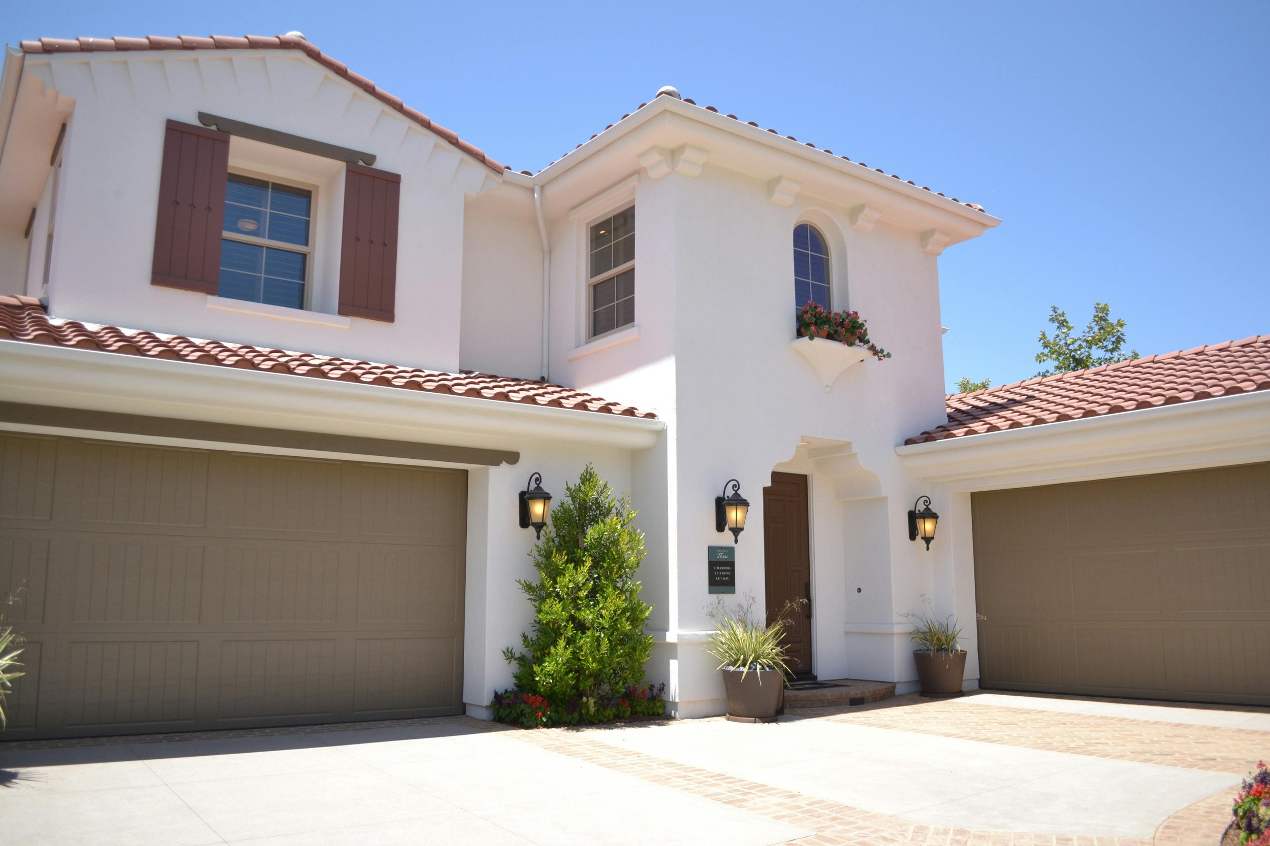 Front view of a modern white house with a brown tile roof, two garage doors, and a small landscaped front yard with green bushes and potted plants. There are three outdoor wall-mounted lamps near the entrance.