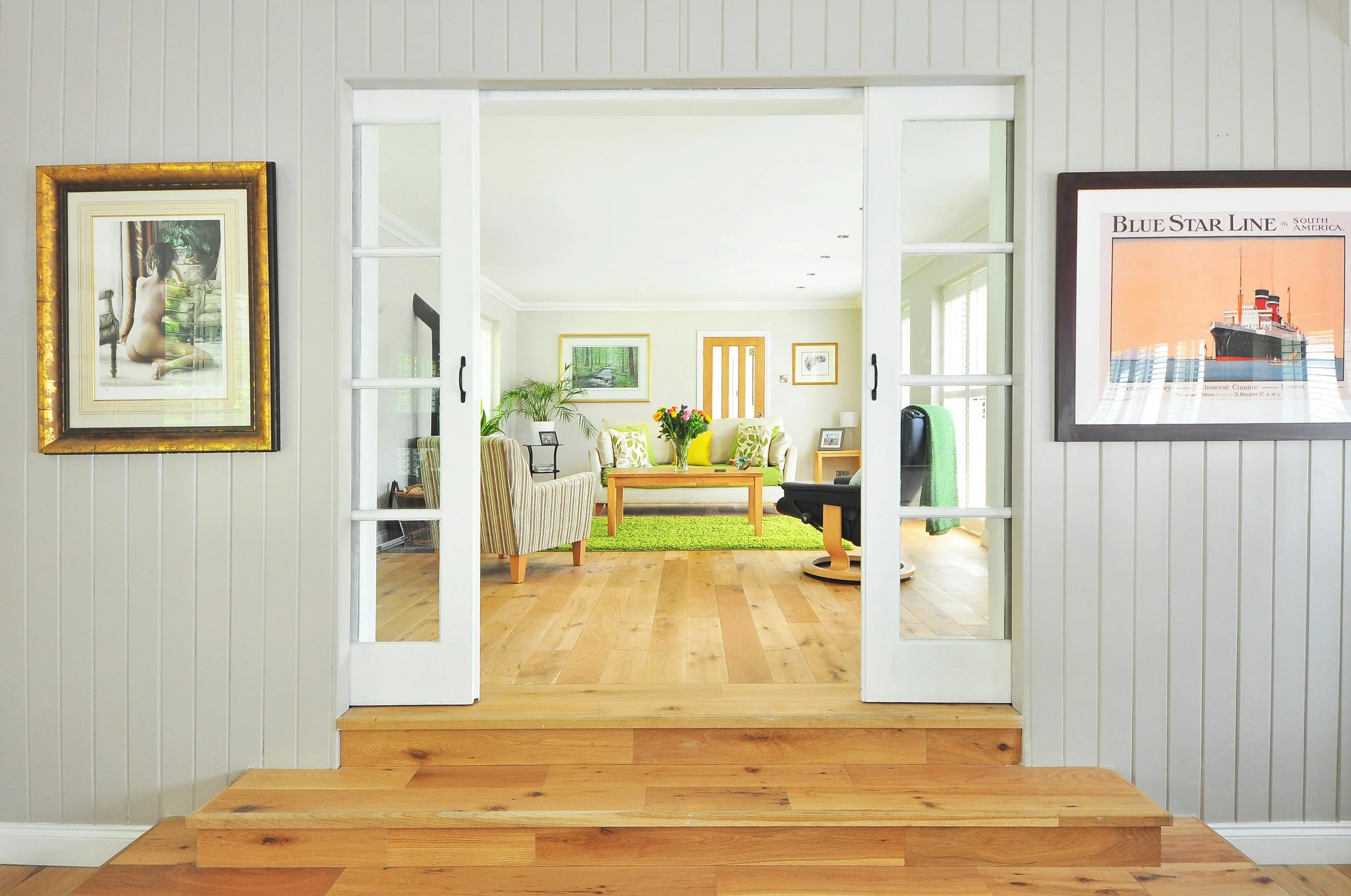 View through a doorway into a living room with light-colored walls, wooden floors, framed artwork, a beige sofa with green and yellow pillows, a wooden coffee table with a flower arrangement, and a green rug. The room is bright with natural light, and there are plants and artwork on the walls.