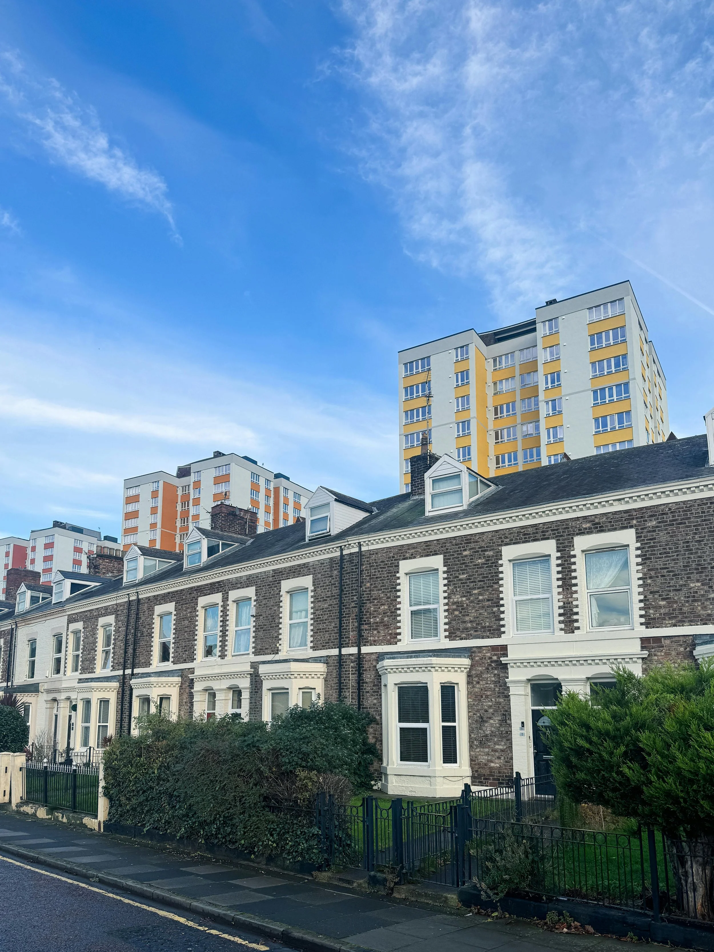 Row of Georgian-style townhouses with brick facades, white trim, bay windows, and small front gardens, with taller modern apartment buildings in the background under a blue sky with some clouds.