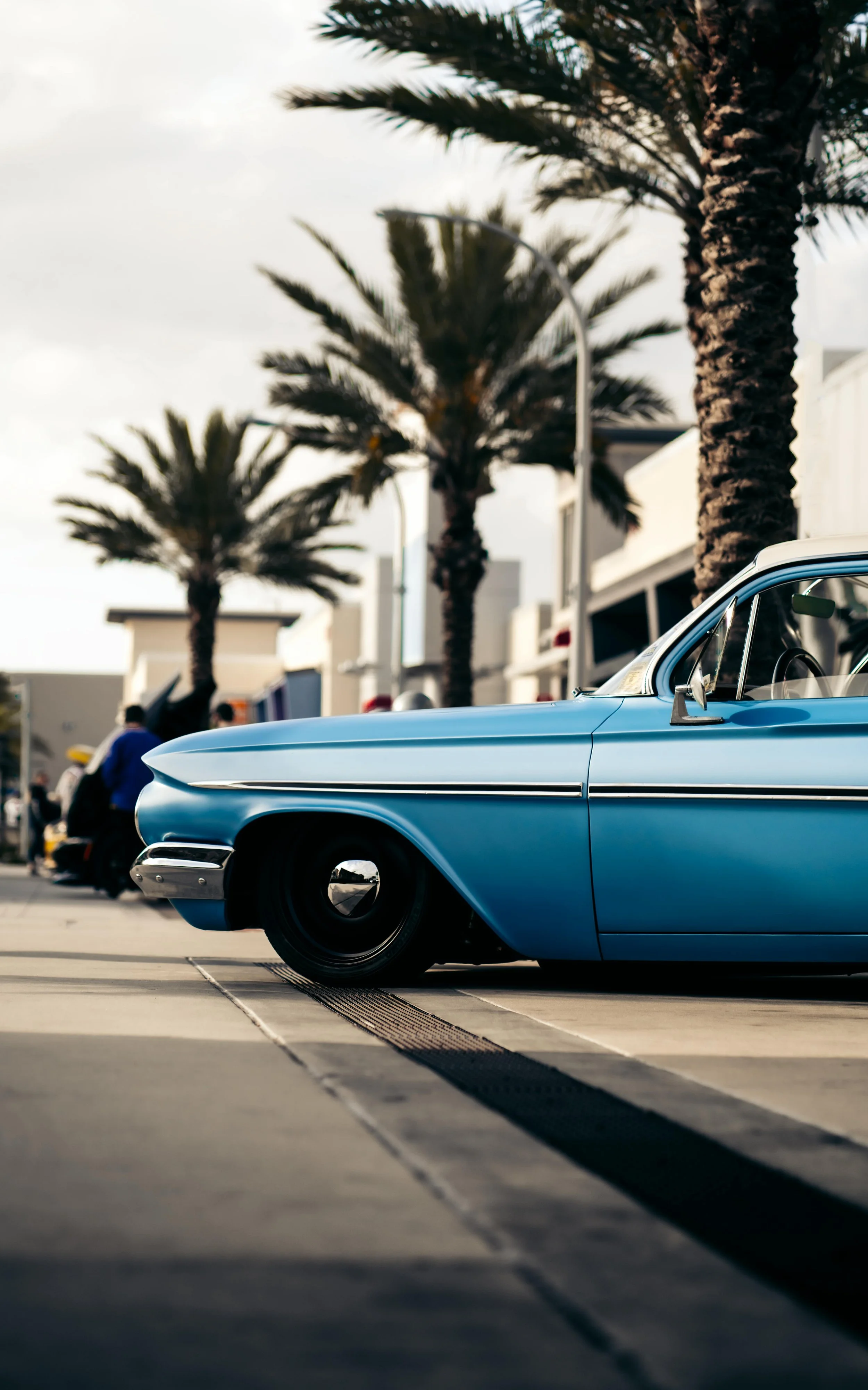A vintage light blue car parked outdoors, with palm trees and a building in the background, and people walking nearby.