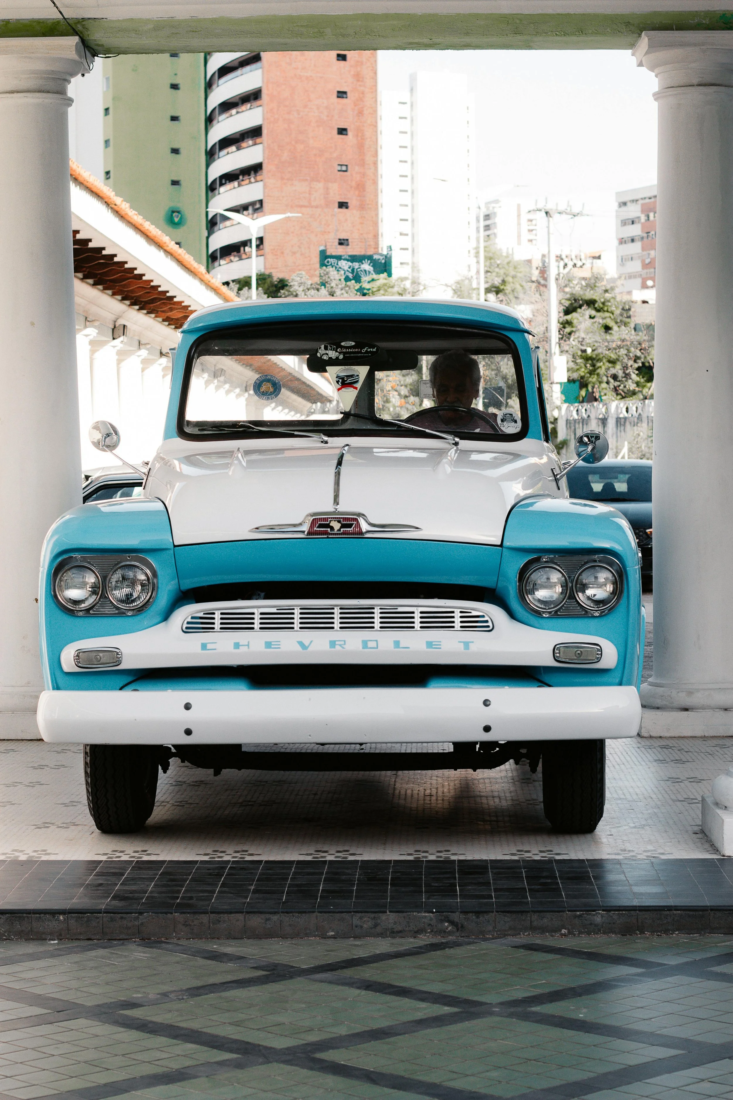 Front view of a vintage Chevrolet truck with a two-tone blue and white paint job, parked under a white archway with columns. Urban buildings are visible in the background.