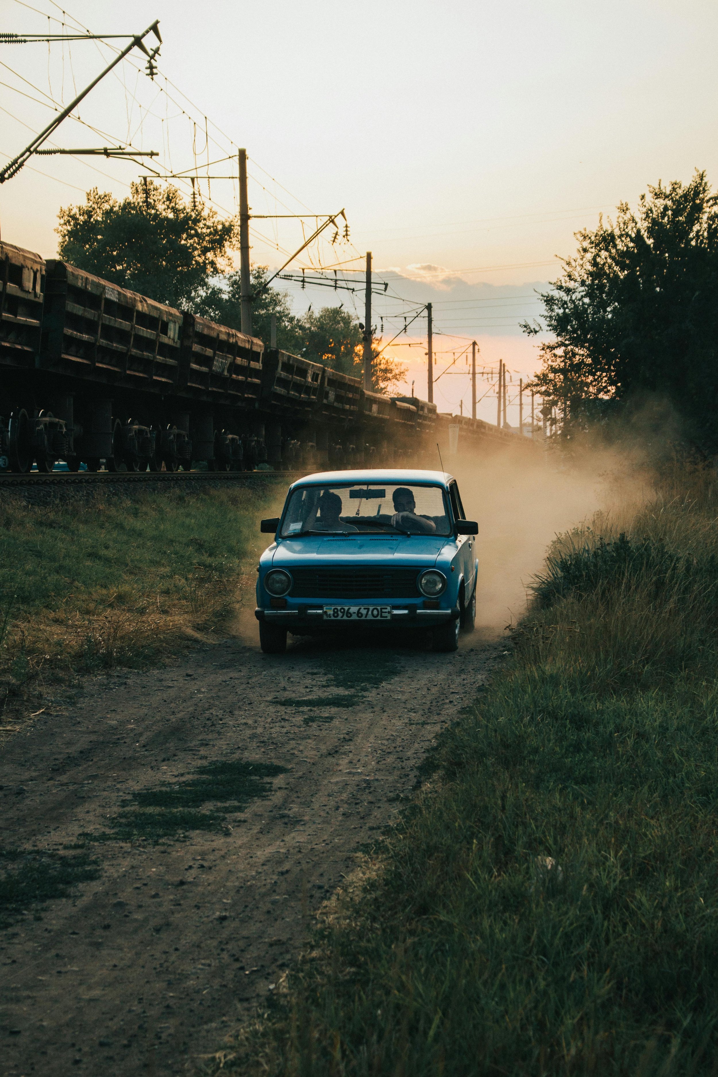 A vintage blue car driving down a dirt road, kicking up dust, with scenery of trees and an electric train passing by on the tracks nearby during sunset.