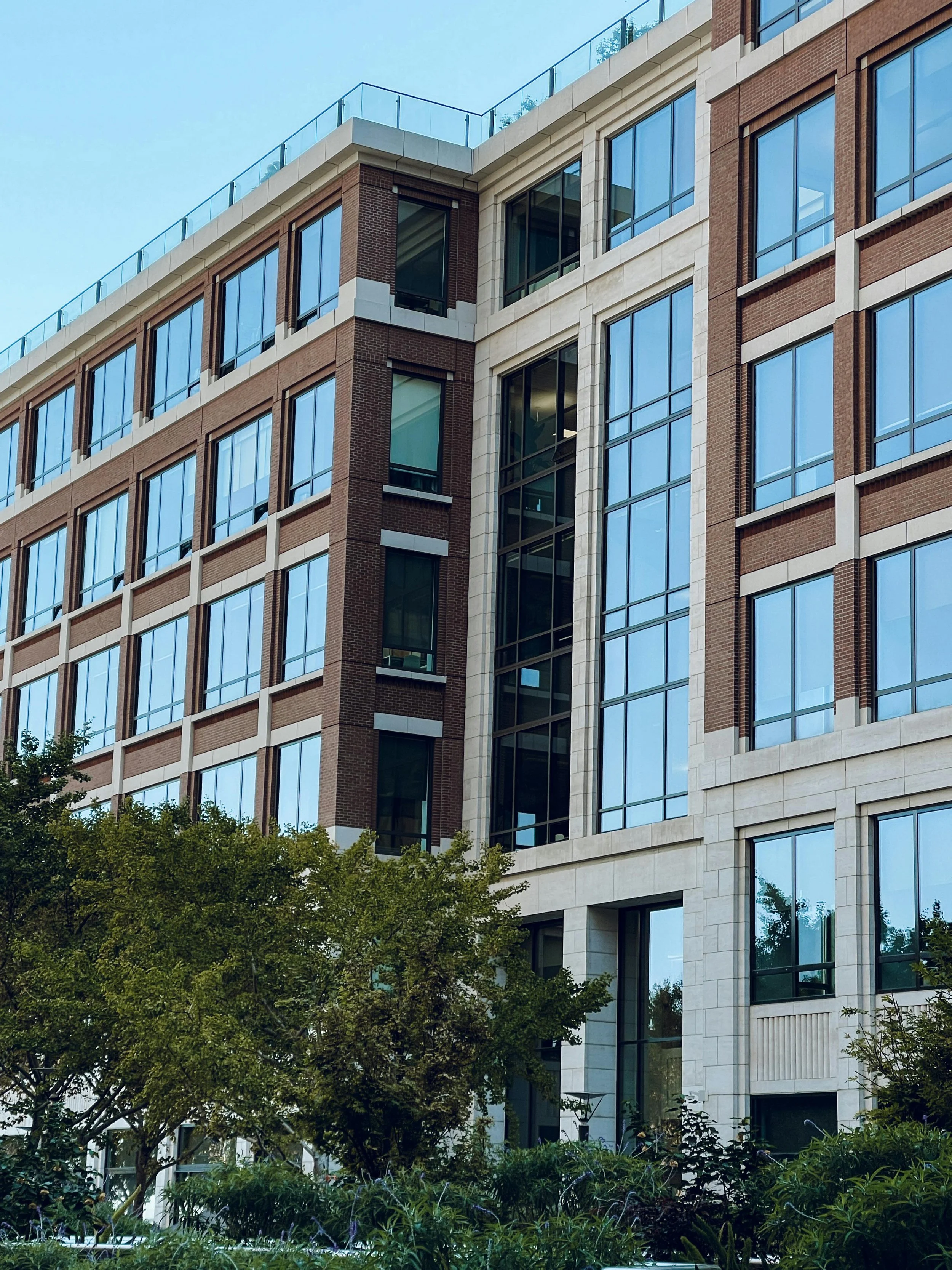 A modern multi-story building made of brick, glass, and stone with numerous large windows. The building is surrounded by lush green trees and shrubs, with a clear blue sky overhead.