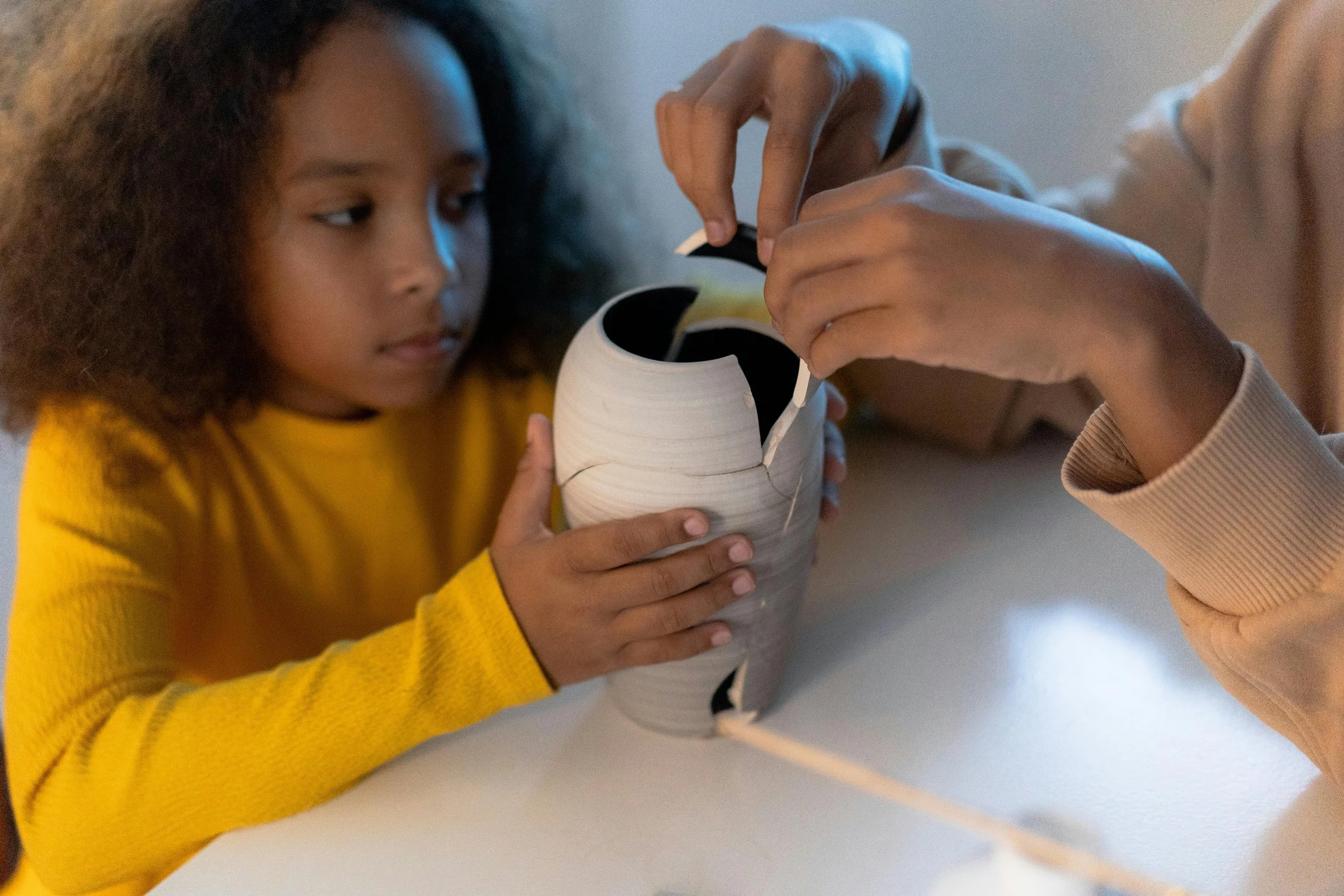 A girl with curly hair wearing a yellow sweater holding a white clay pot with piece missing, the pot has a black interior and a white design, and another person, wearing a beige top, pute the missing piece of the pot back