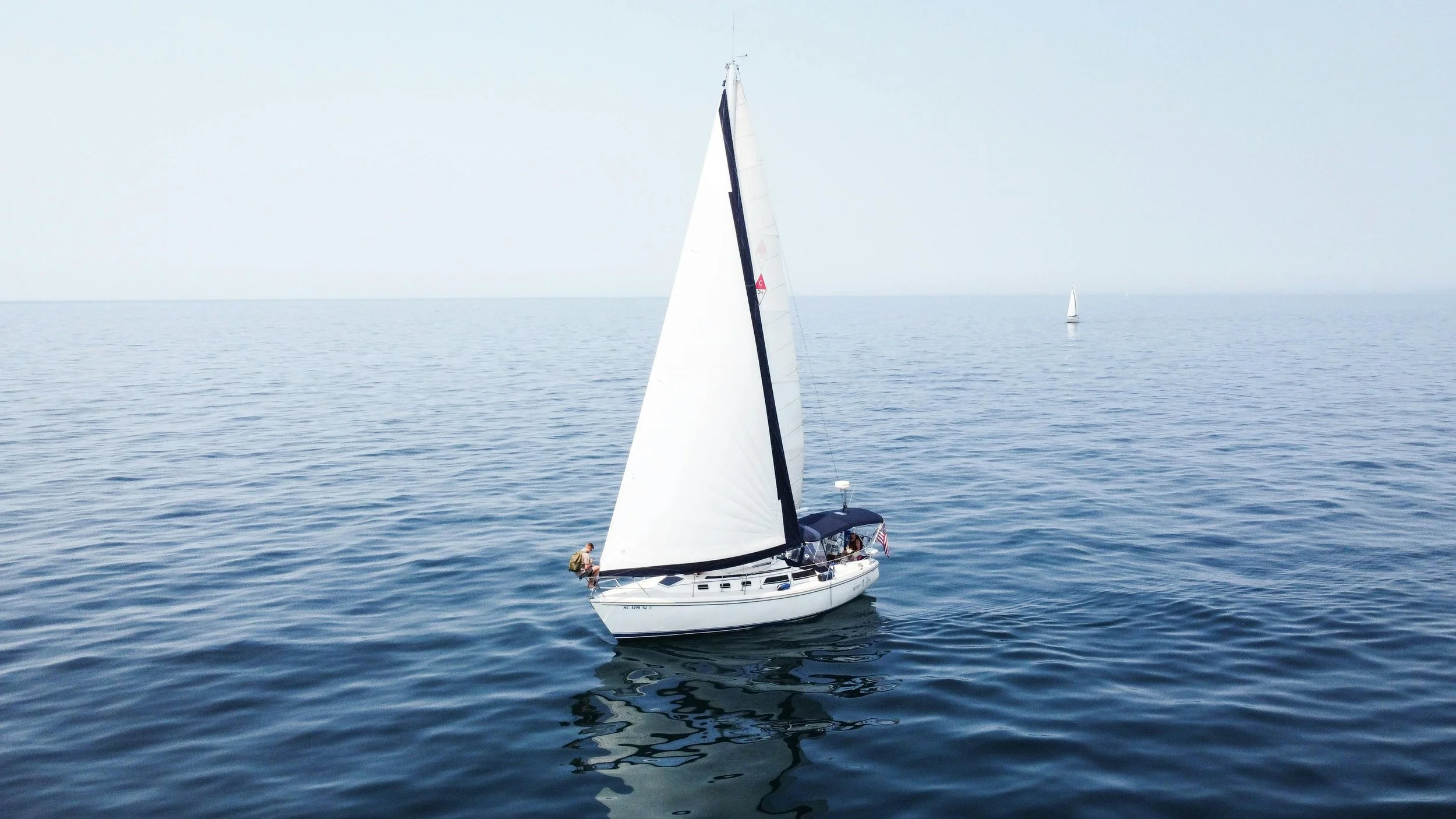 A white sailboat on calm blue water with a large sail, with two people on the boat and another sailboat in the distance. boat insurance coverage offered by Hardin Insurance