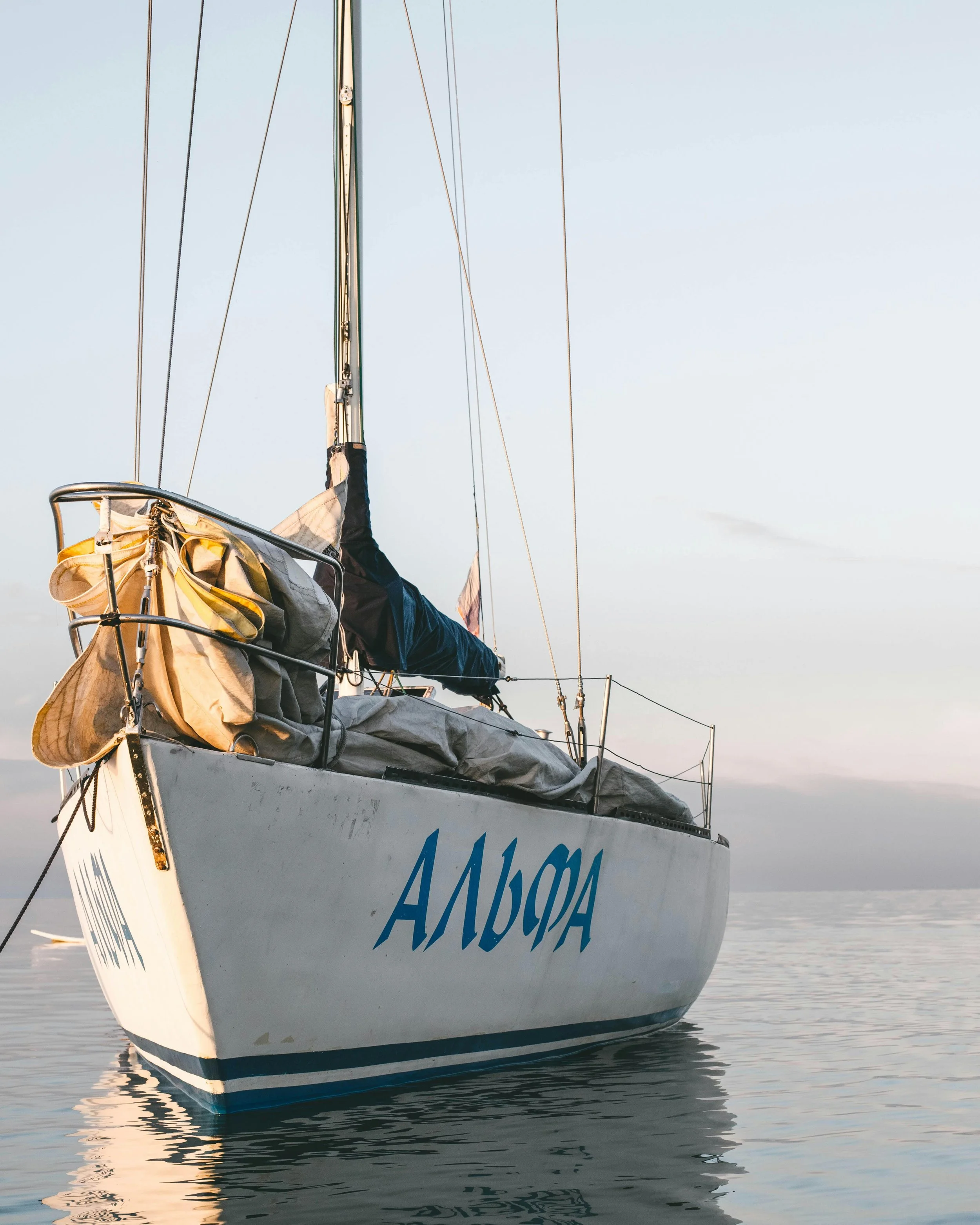 Close-up of the bow of a sailboat with the registration number A1464A, floating on calm sea waters during sunset or sunrise, with a light-colored sky and gentle reflections in the water.