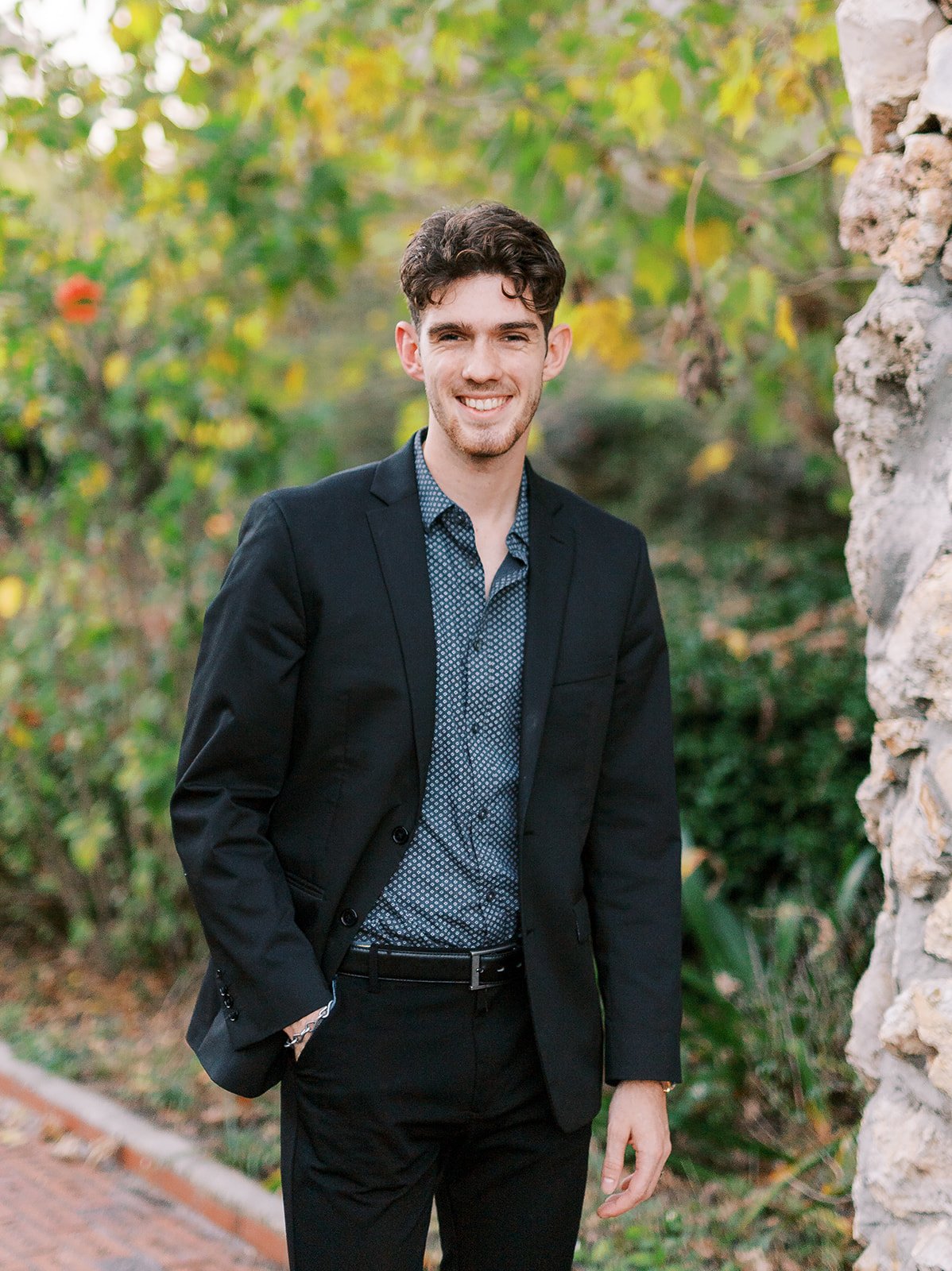 John Hardin  Agent with Hardin Insurance wearing a dark suit jacket over a patterned shirt, standing outdoors with greenery and a stone wall in the background.