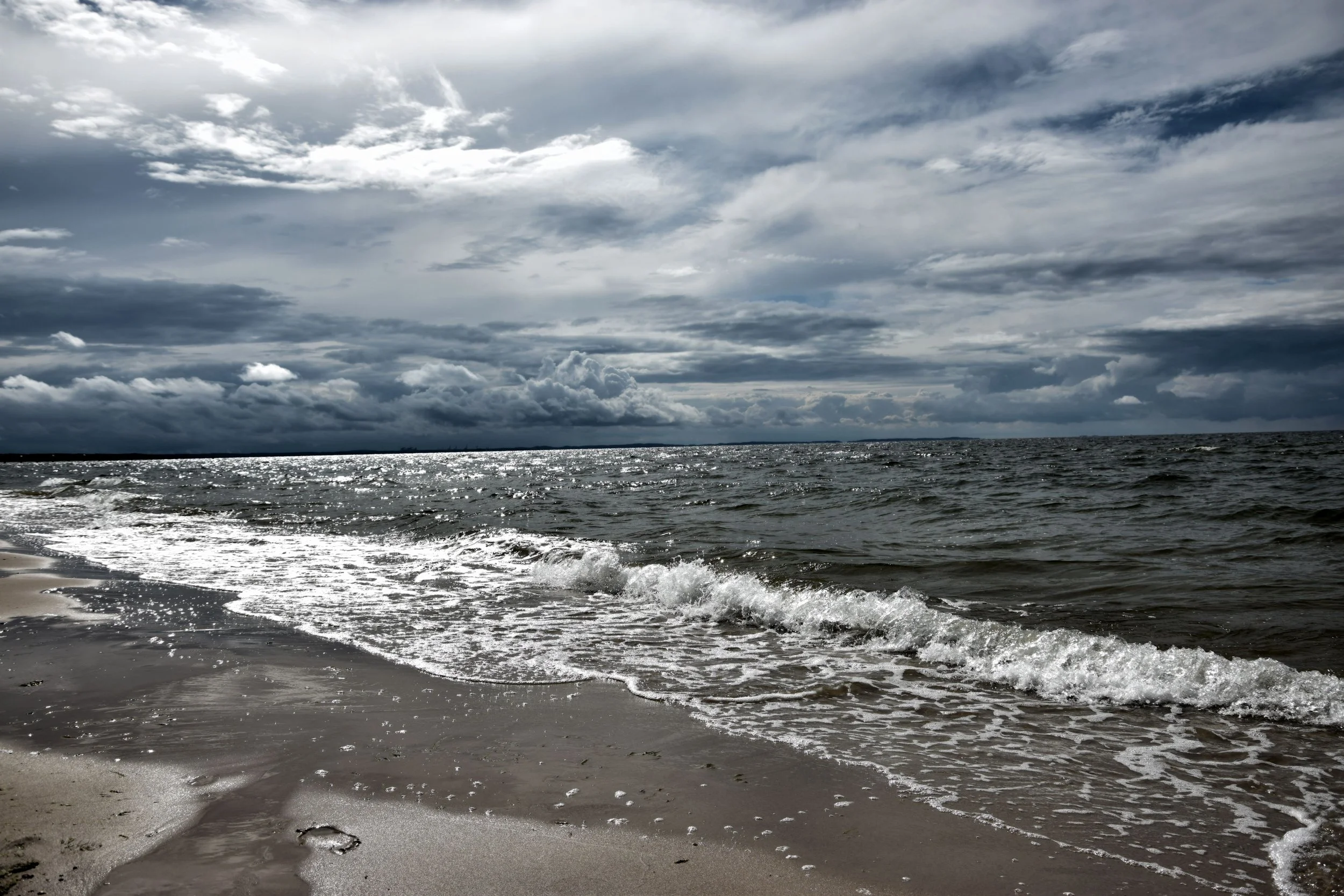 Overcast sky with dark clouds over the ocean, gentle waves lapping a sandy beach with footprints.