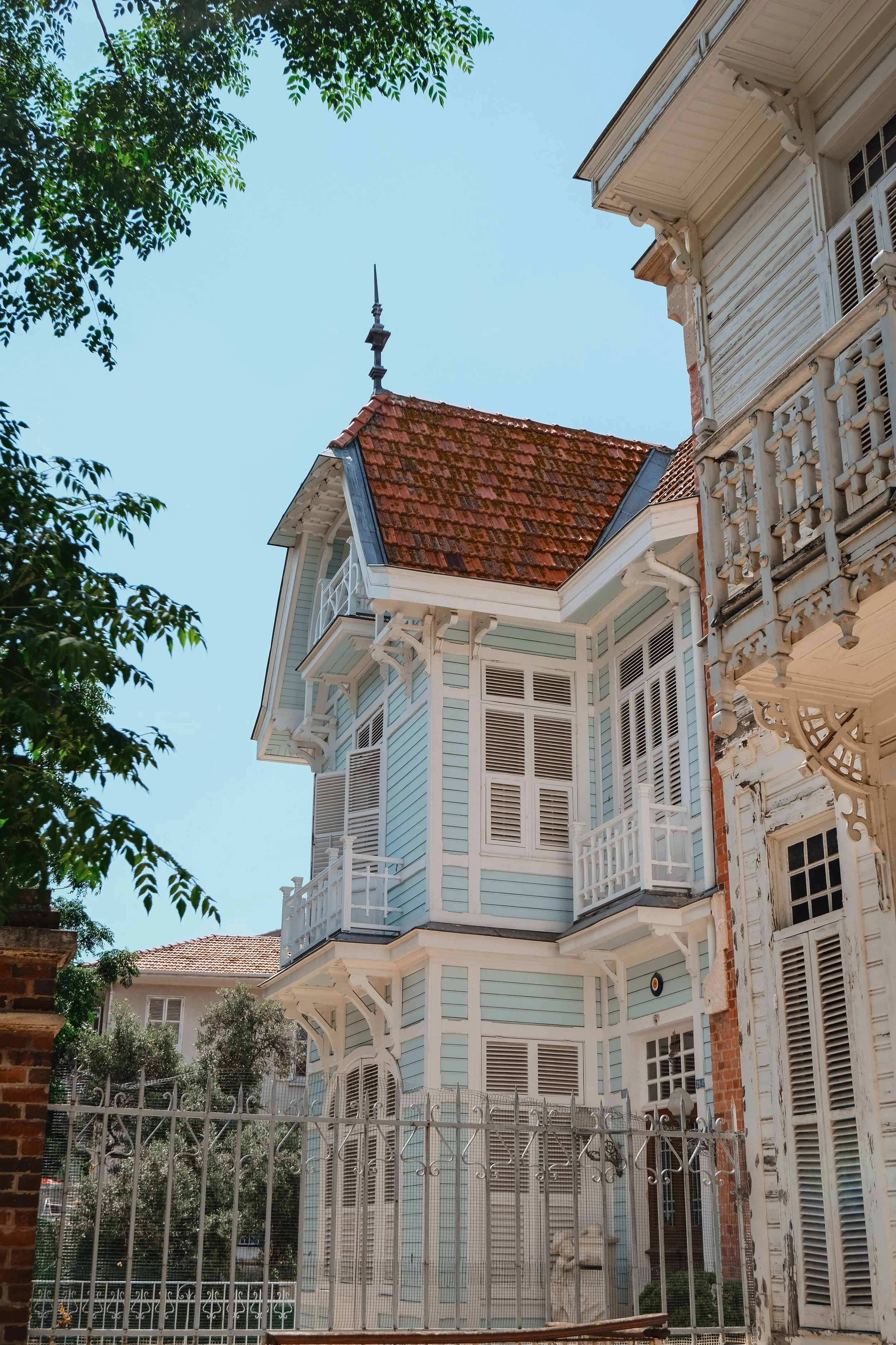 Three-story Victorian-style house with light blue exterior, ornate trim, multiple balconies, and a red-tiled roof. The house is surrounded by a metal fence, and there are trees in the background and foreground.