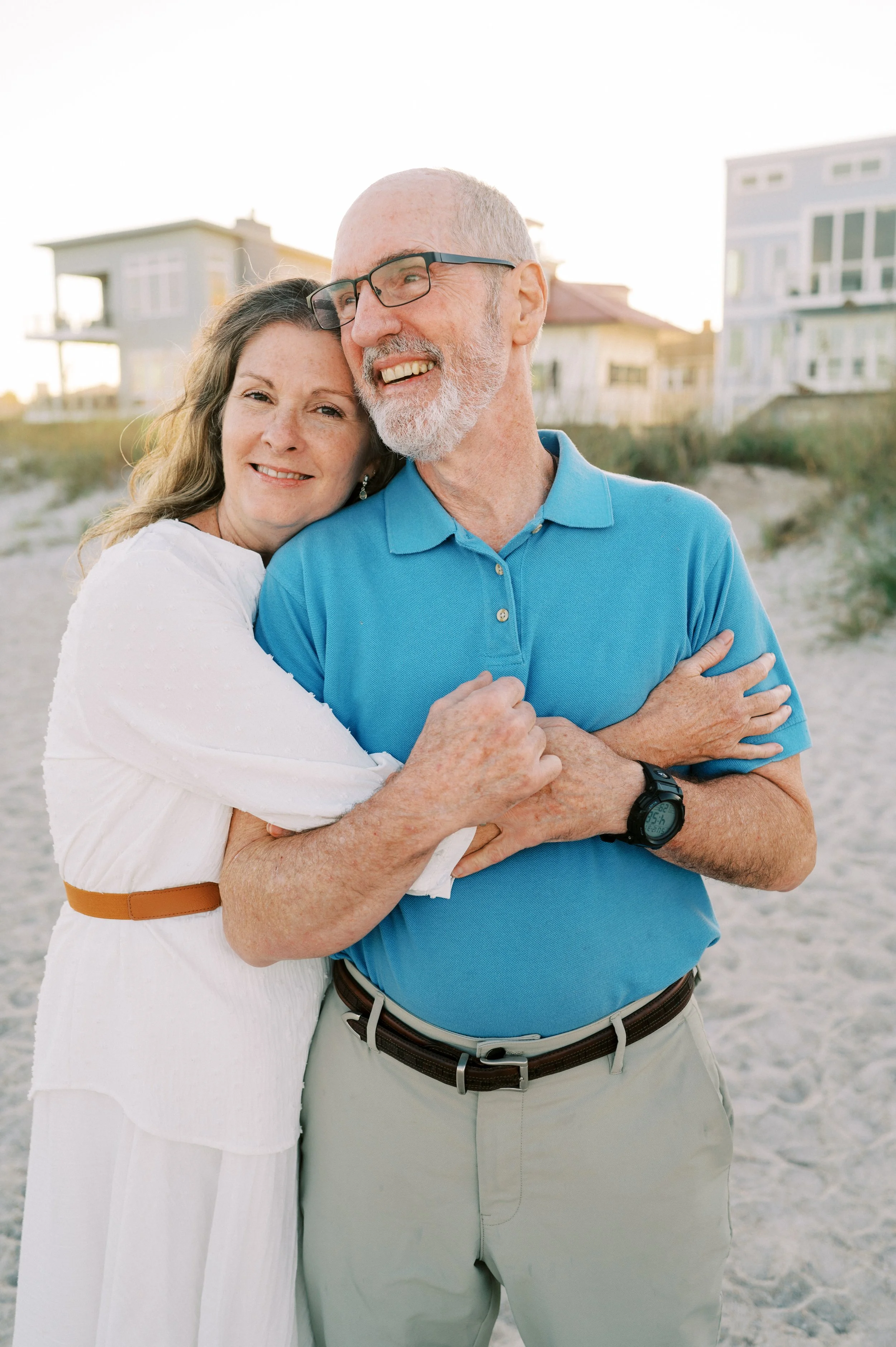 Michael and Tracy Hardin smiling and hugging on Fernandina Beach with houses in the background during sunset.