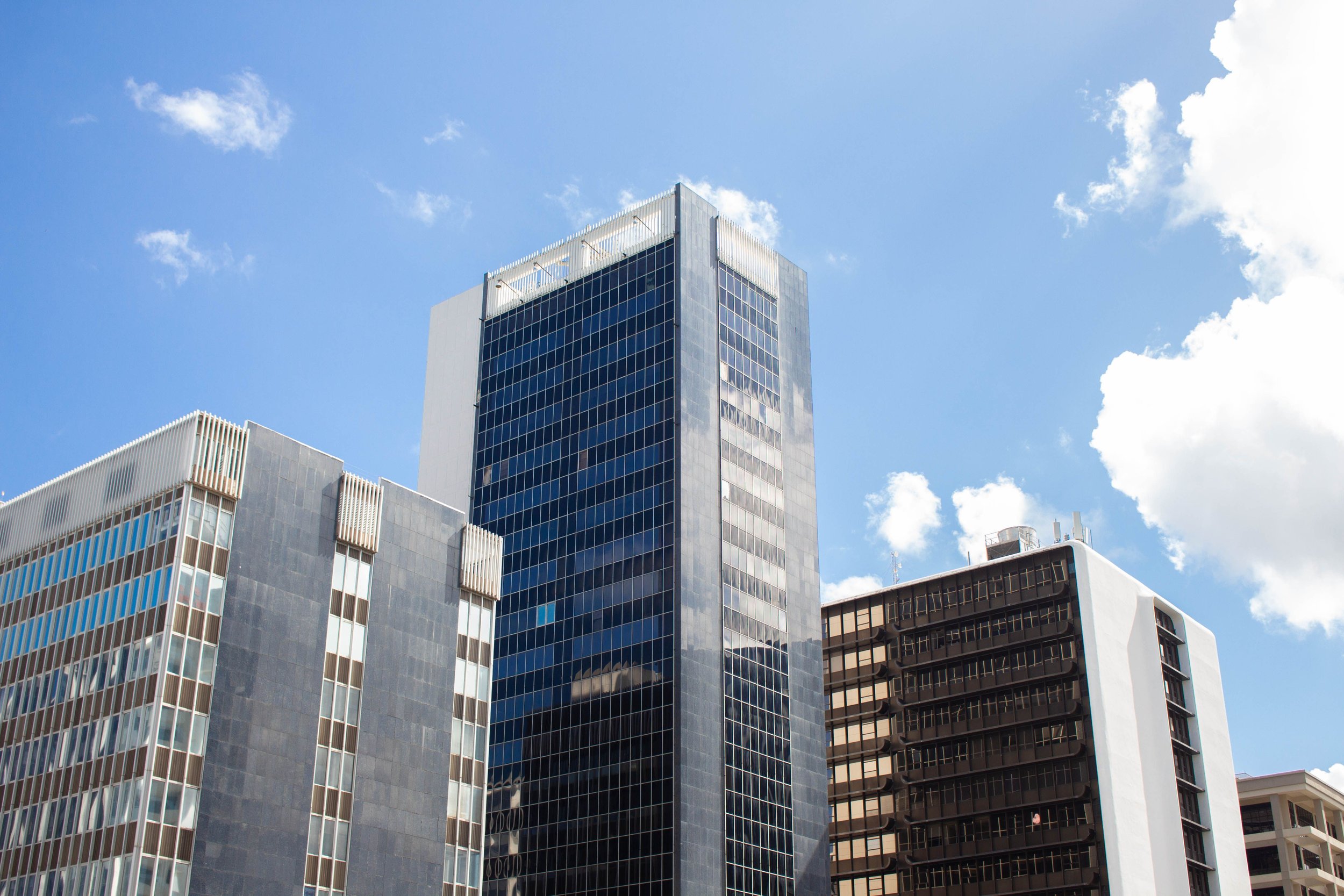 A cityscape featuring several modern tall buildings with glass facades under a bright blue sky with scattered clouds.