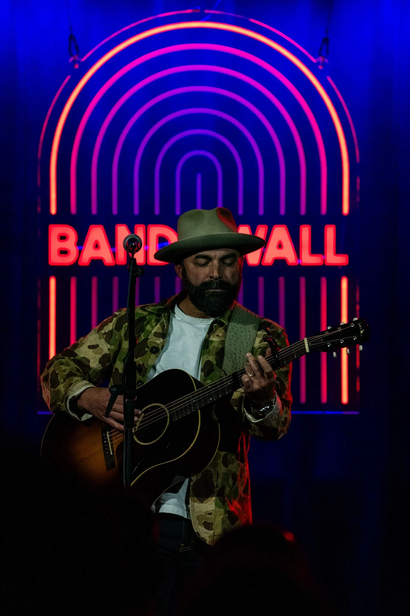 Drew Holcomb playing a concert at Band on the Wall in Manchester, England. A man playing guitar on stage in front of neon lights.