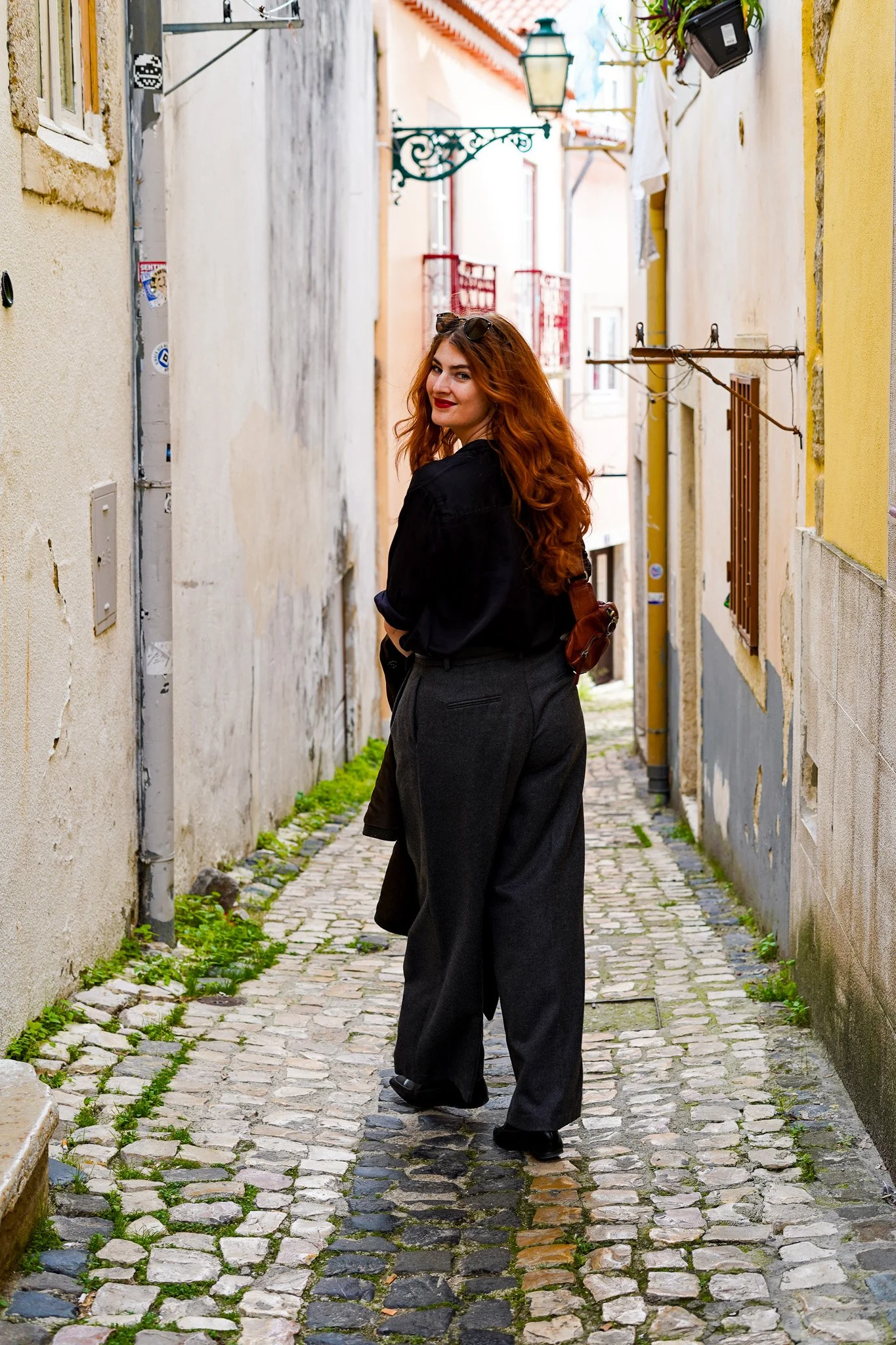 Well dressed woman with red hair looks back to the camera as she is walking down a cobble stone street in Lisbon, Portugal