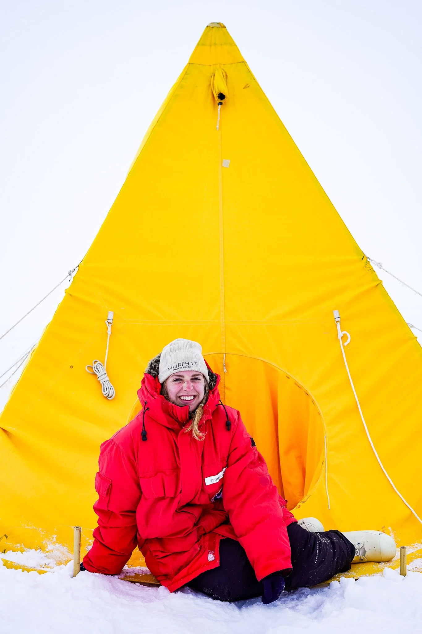Woman in large red parka sitting on the snow in front of yellow tent on Mt. Erebus, Ross Island, Antarctica