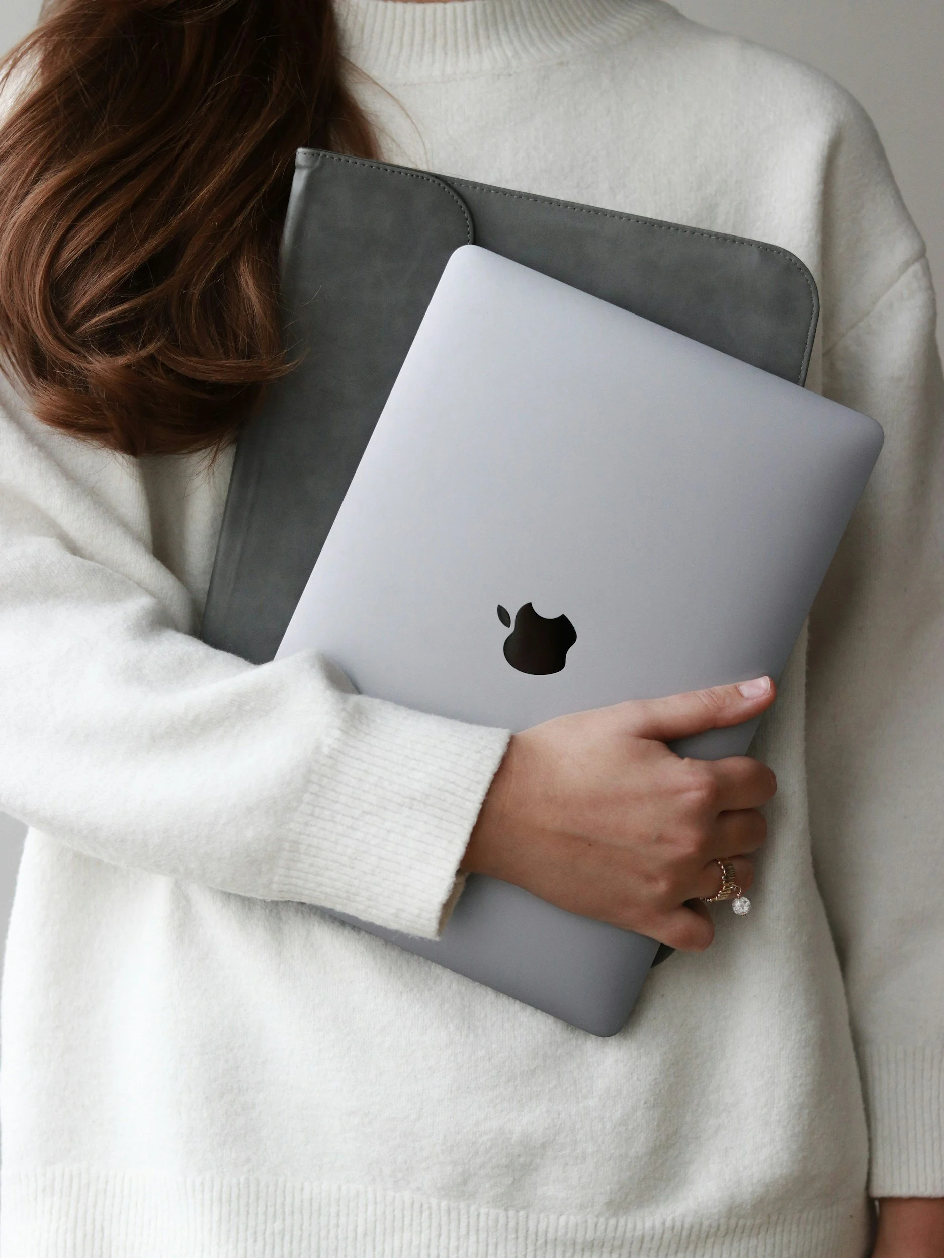 A person wearing a white sweater holding a silver MacBook laptop close to their chest.