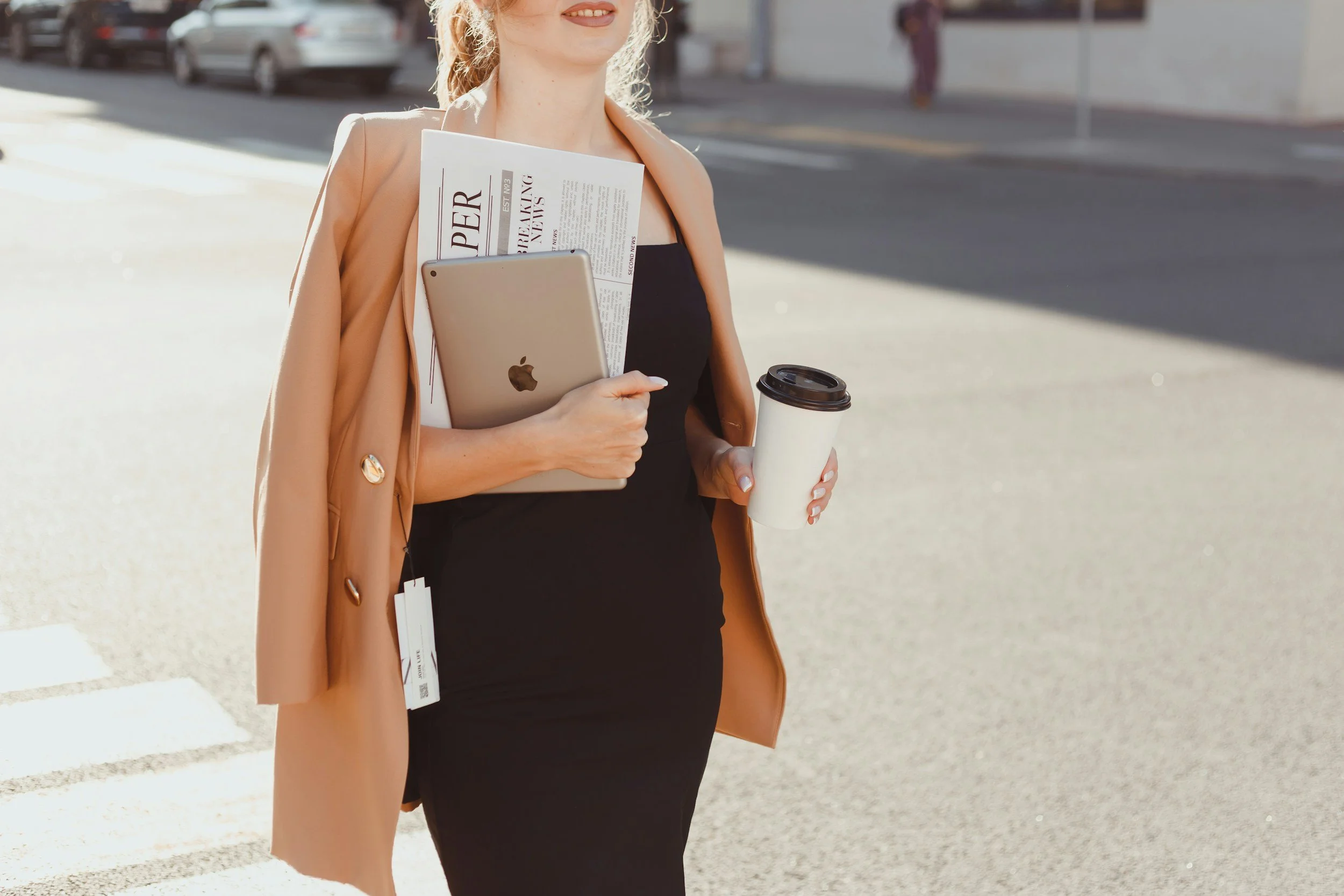 A woman walking outside holding a coffee cup, an iPad, and newspapers, wearing a black dress and a tan blazer, in an urban area.