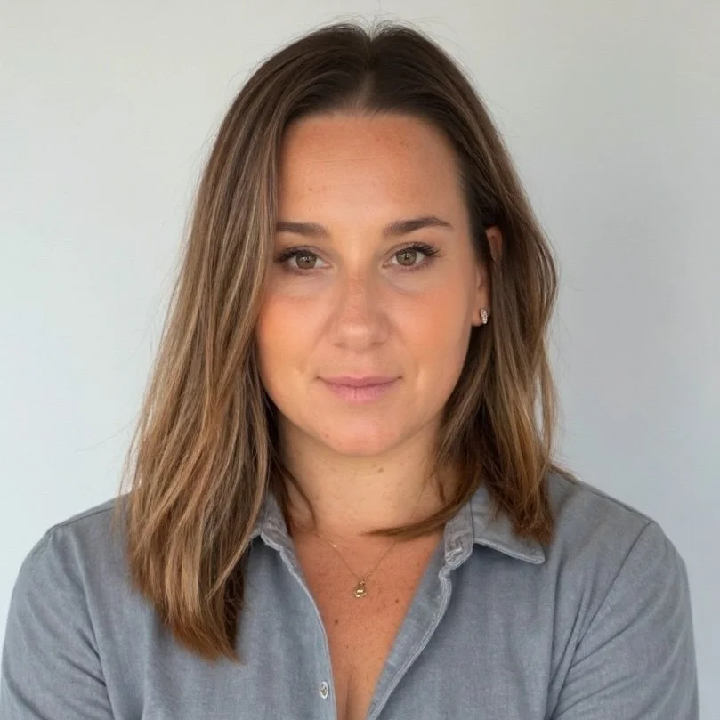 A woman with shoulder-length brown hair wearing a gray button-up shirt and gold necklace, posing in front of a plain light-colored background.
