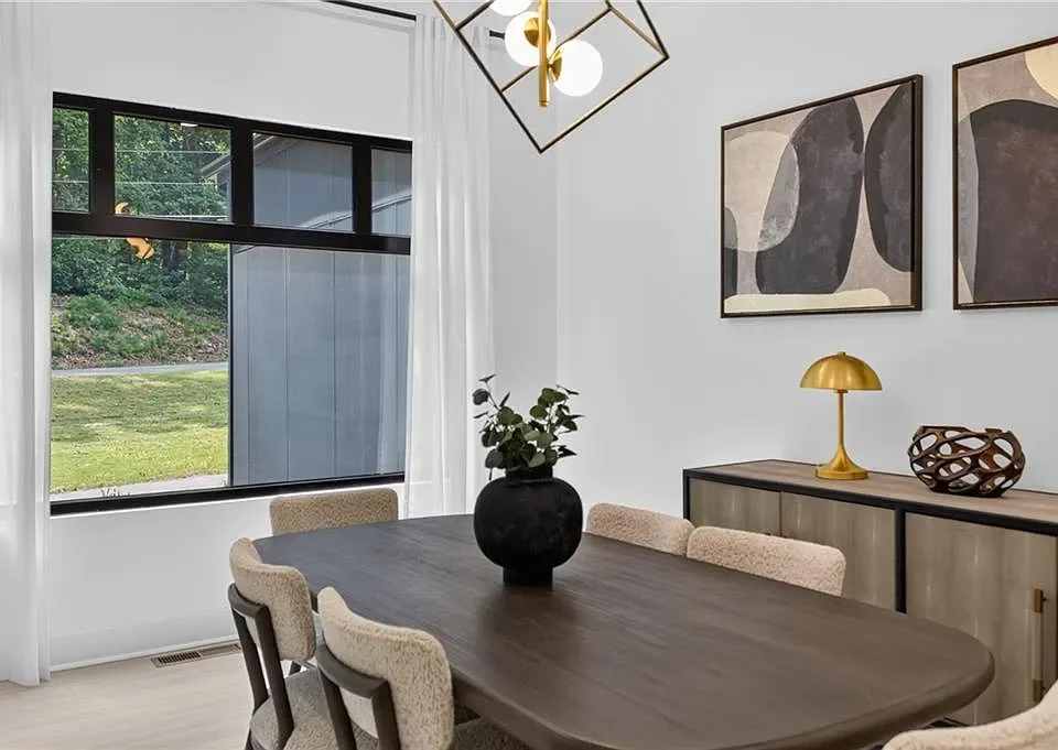 A dining room with a dark wood table, beige upholstered chairs, a black vase with greenery, abstract artwork on the wall, a gold table lamp, decorative bowl, and large window with white curtains showing an outdoor yard.