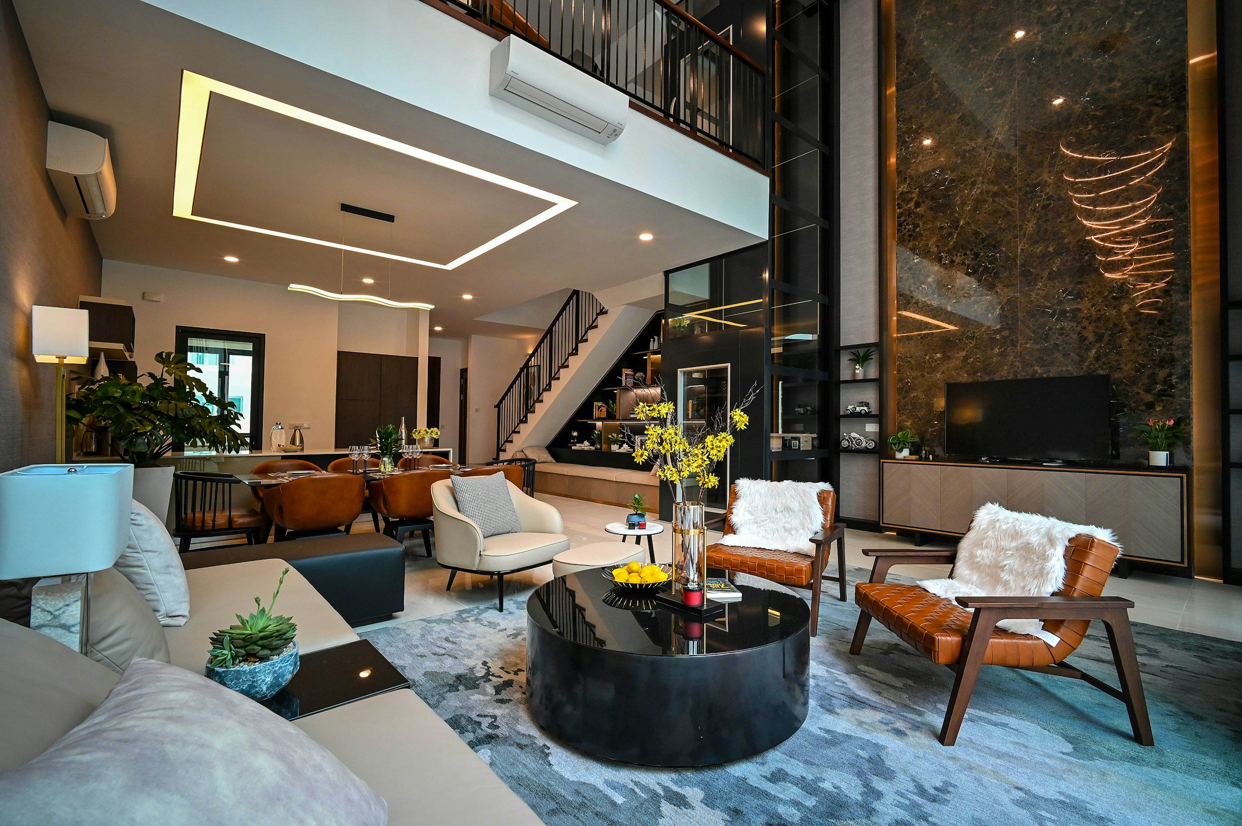 Modern living room with tan, beige, and brown furniture, including armchairs and a sofa, decorated with pillows and throw blankets, a black round coffee table, and various plants, set in a spacious area with a high ceiling, wooden staircase, and decorative lighting fixtures.