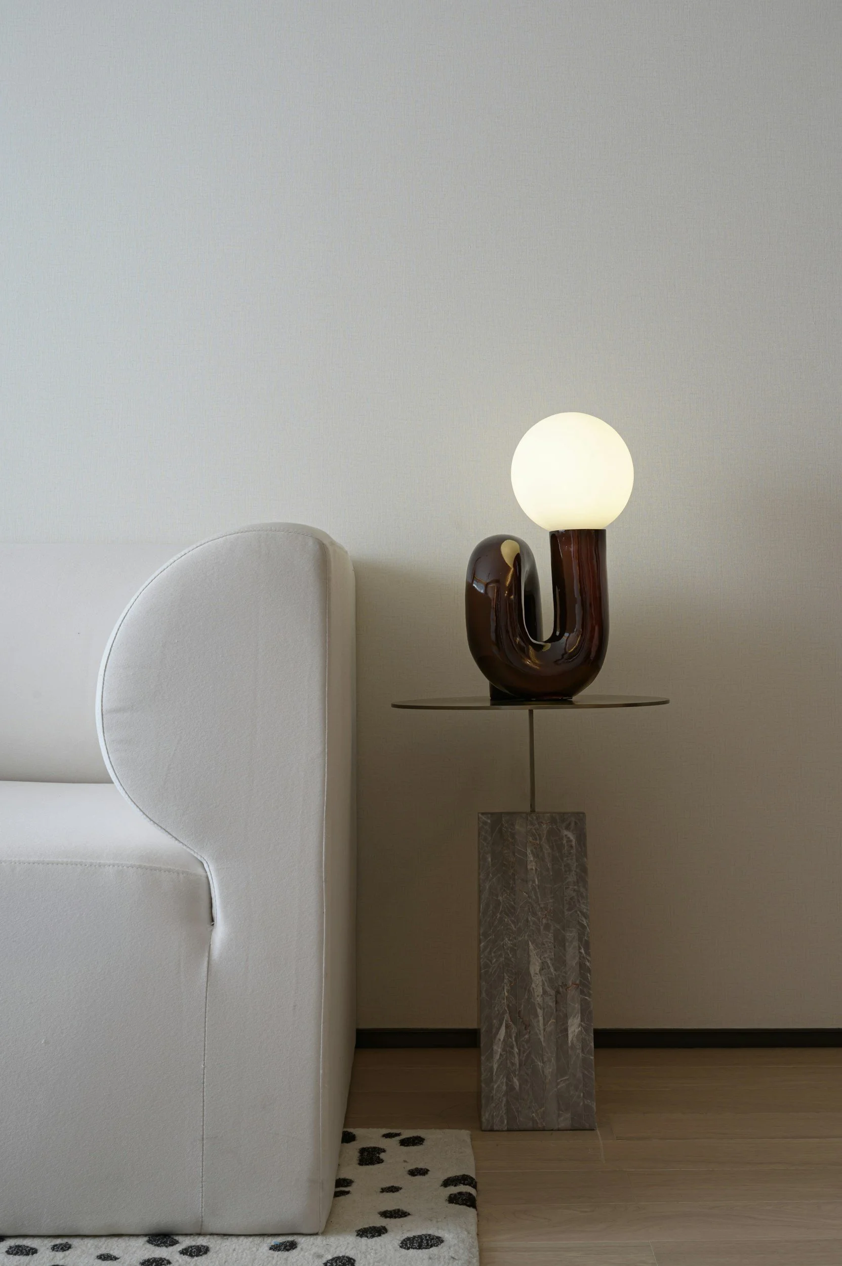 Interior corner with a white armchair on a black and white polka dot rug, a marble pedestal, a modern abstract lamp with a frosted globe and a dark brown base, and a beige wall.