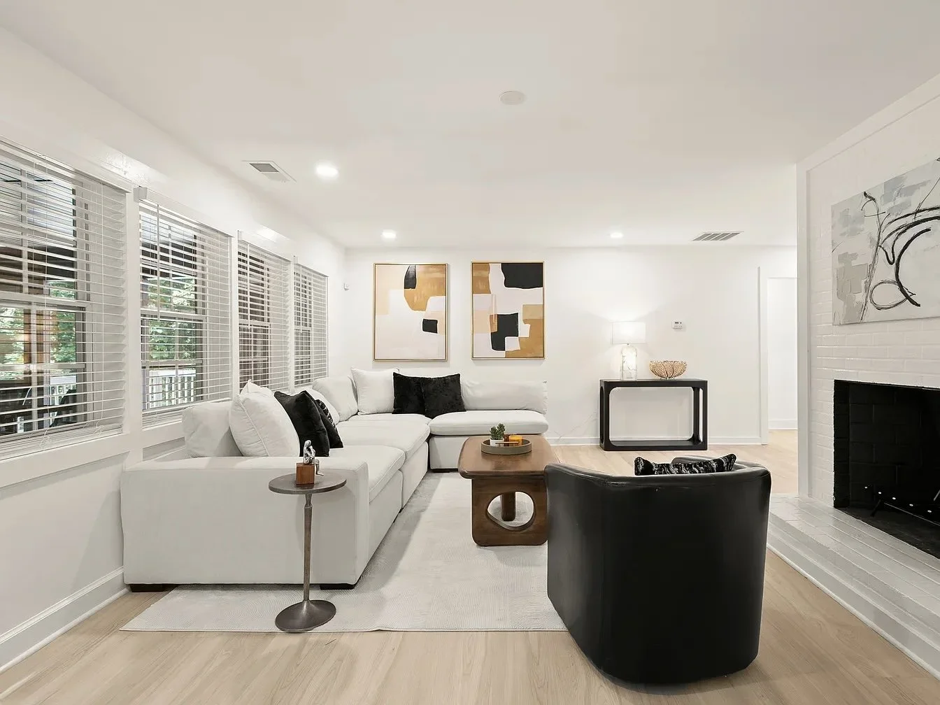 Bright living room with white sectional sofa, black and white pillows, abstract wall art, wooden coffee table, black armchair, white painted brick fireplace, and large windows with white blinds.