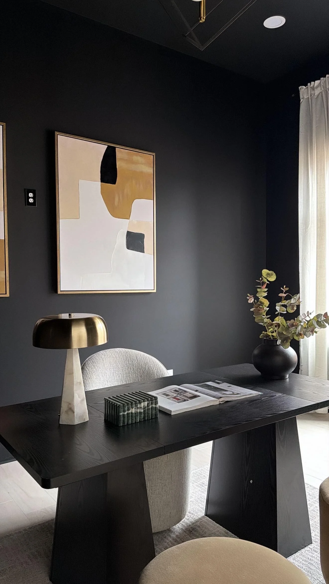 A modern dining room with a dark wall, abstract artwork, a black dining table, beige chairs, a metallic table lamp, a black vase with greenery, and a window with white curtains.