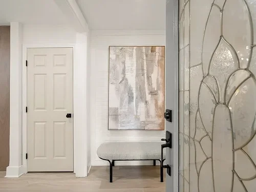 Interior view through a partially open door with stained glass, showing a hallway with a white door, a large abstract painting, and a bench.