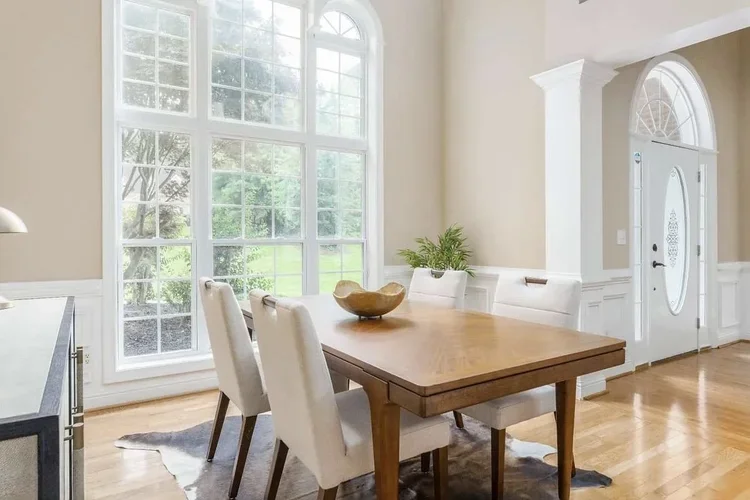 Bright dining area with large windows, a wooden table with four upholstered chairs, a decorative bowl, and a sideboard with a plant, near an entry door with an arched window.