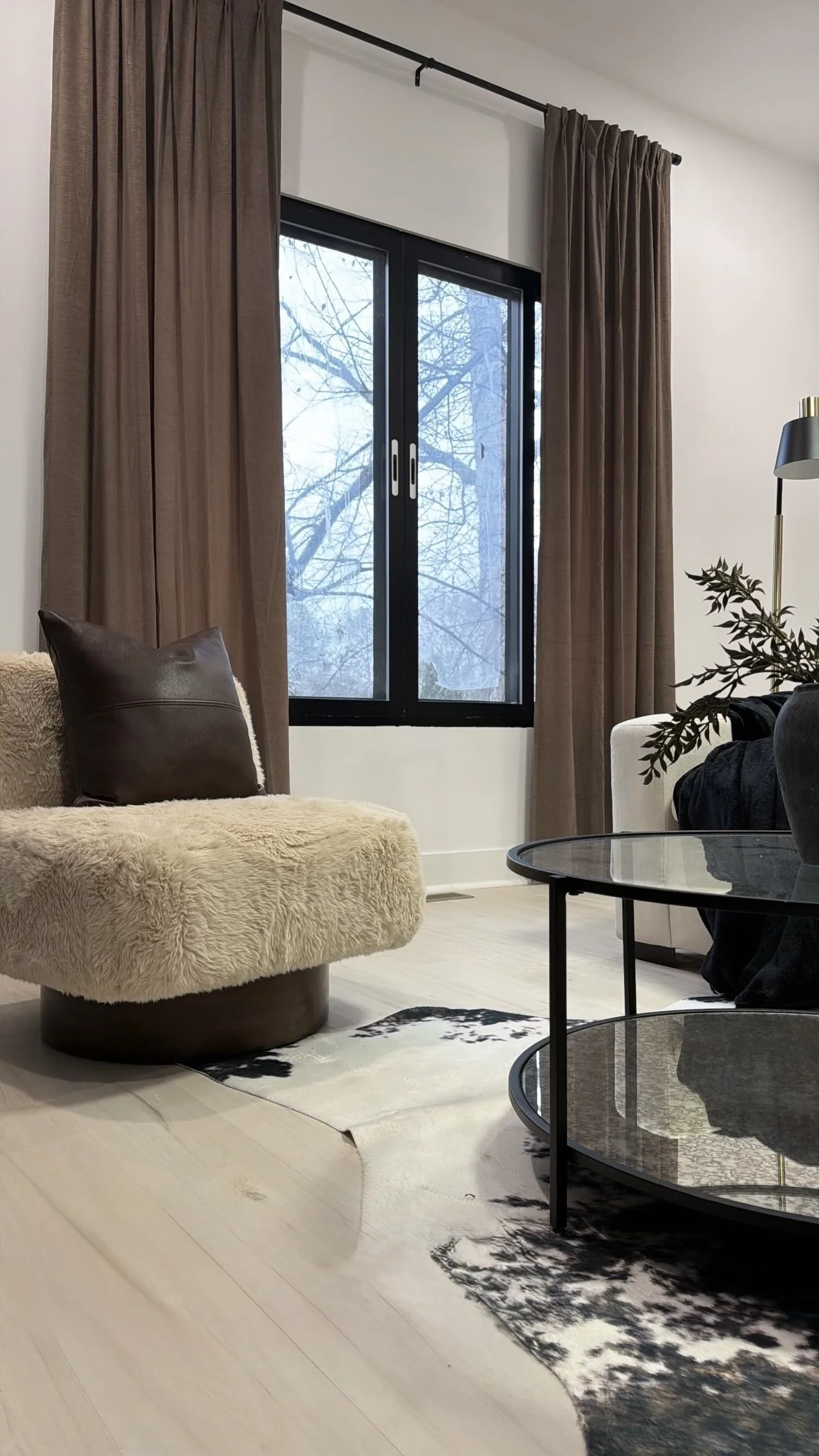 Living room with a window, brown curtains, a beige plush chair with a dark brown pillow, a black and gold standing lamp, a glass coffee table, a decorative plant, and a black and white patterned rug.