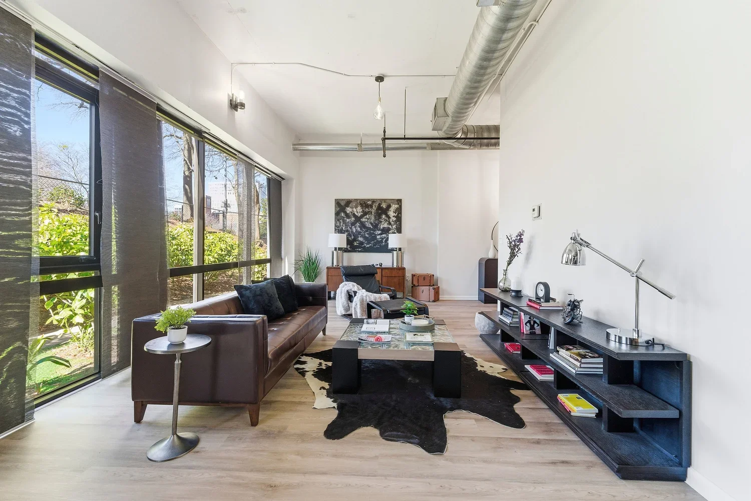 Modern living room with large windows, brown leather sofa, black and white cowhide rug, black wooden TV stand with books and decorations, glass coffee table, armchair with throw blanket, side table with potted plant, and contemporary artwork on the w
