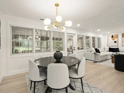 Living room with large windows, a round dining table with six white chairs, a modern chandelier, and a white sofa.