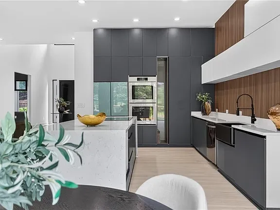 Modern kitchen with black and white cabinets, a marble island, and kitchen appliances including a built-in oven and a refrigerator, with wooden accents and decorative plants.