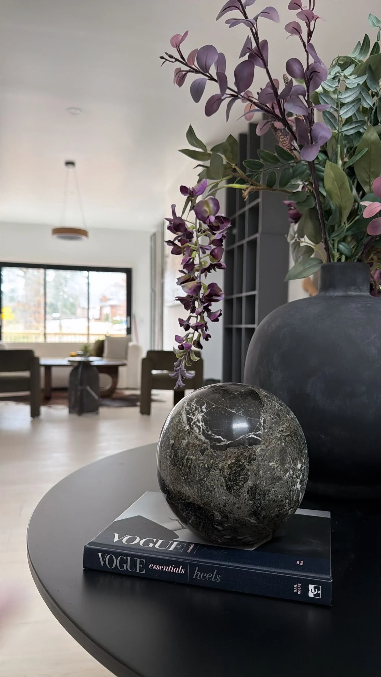 A round table with two decorative stones and a vase with purple flowers, resting on a Vogue magazine, in a modern living room with large windows and minimal decor.