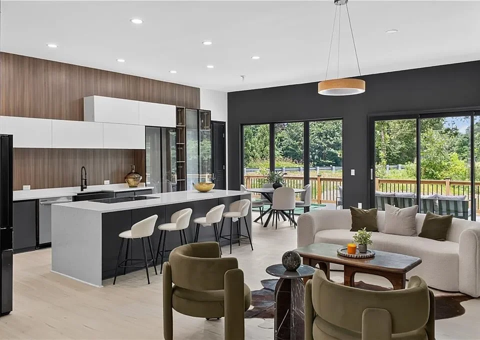 Modern open-plan living room and kitchen with white and black cabinetry, a white kitchen island with four barstools, a white sofa with pillows, a wooden coffee table, armchairs, large windows, and a sliding glass door leading to a deck with outdoor f