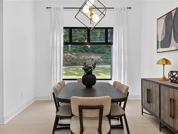 Modern dining room with a dark wood table, six beige upholstered chairs, a large window with white curtains, a black vase with flowers, and a sideboard with a gold table lamp and artwork on the wall.