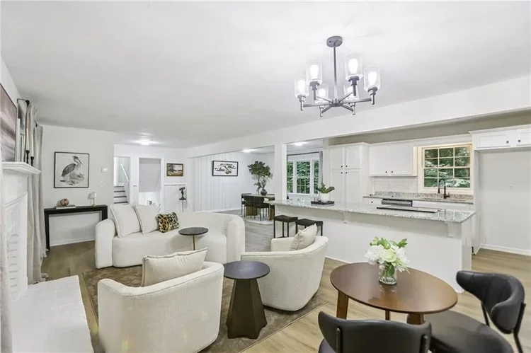 Spacious living room with white sofa and armchairs, black side tables, a round dining table with a floral arrangement, open kitchen with white cabinets, granite countertops, and windows with greenery outside, modern chandelier, hardwood floors.