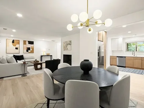 Open-concept living and dining area with a black round table, white upholstered chairs, modern chandelier, white walls with abstract art, fireplace, and kitchen with wooden cabinets.