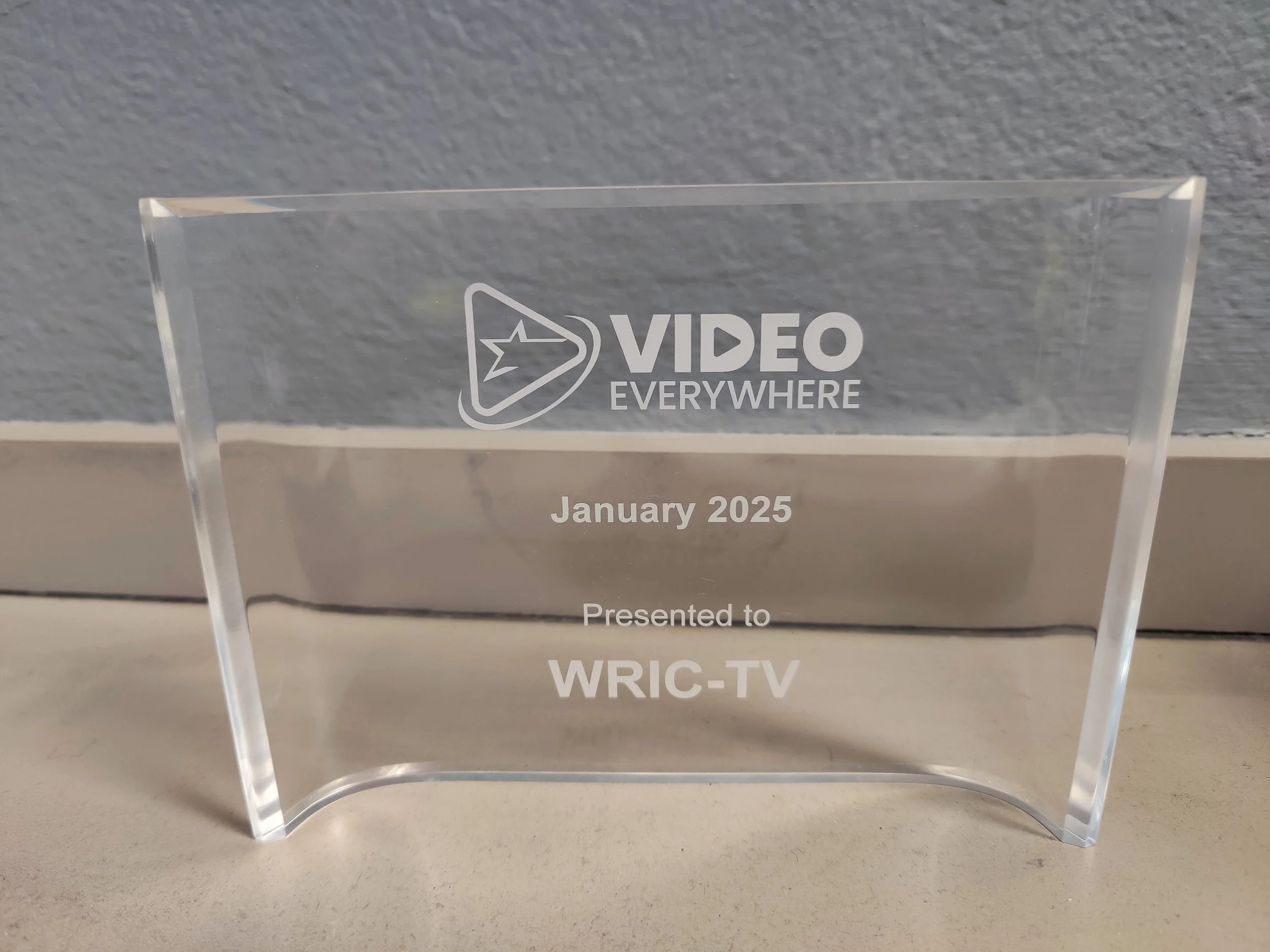 A clear acrylic award plaque with logo and text, including words 'Video Everywhere,' 'January 2025,' and 'Presented to WRIC-TV,' positioned against a gray textured wall.