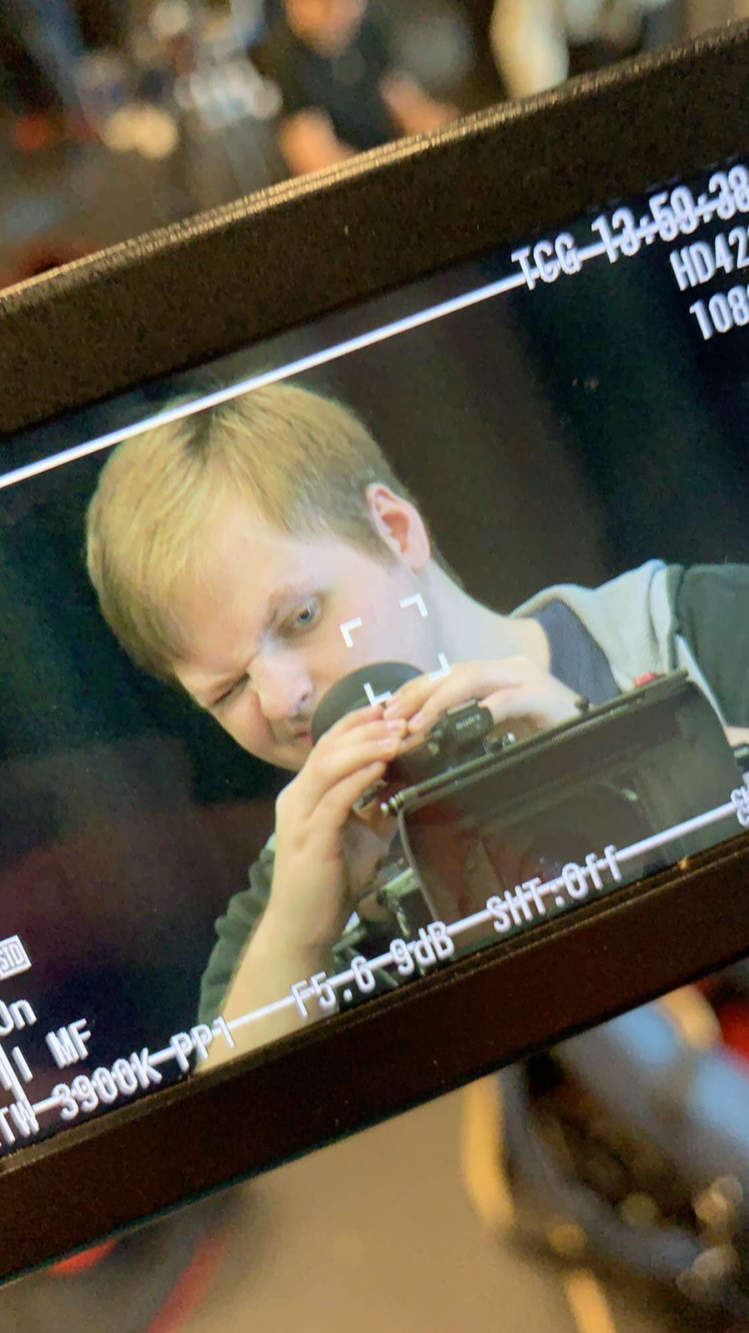 A young man with blond hair looking through a camera viewfinder and holding the camera with both hands, winking with one eye closed.