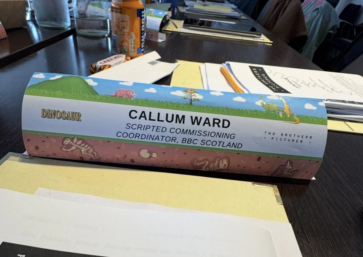 Nameplate on desk identifying Callum Ward as scripted commissioning coordinator for BBC Scotland, with colorful cartoon dinosaur and landscape illustrations.