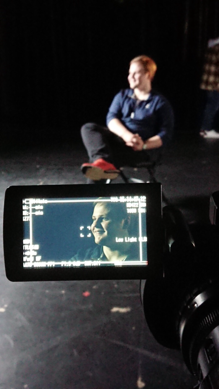 A person sitting on a chair on a dark stage, being filmed or photographed, with the camera's display showing a smiling woman with blonde hair.