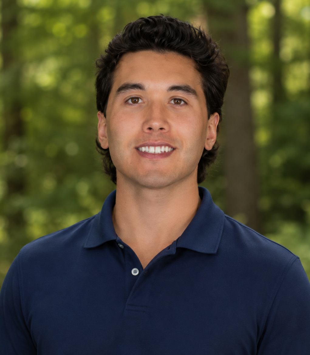 Portrait of a young man with dark hair and a friendly smile, wearing a navy polo shirt, standing outdoors in a lush green forest.