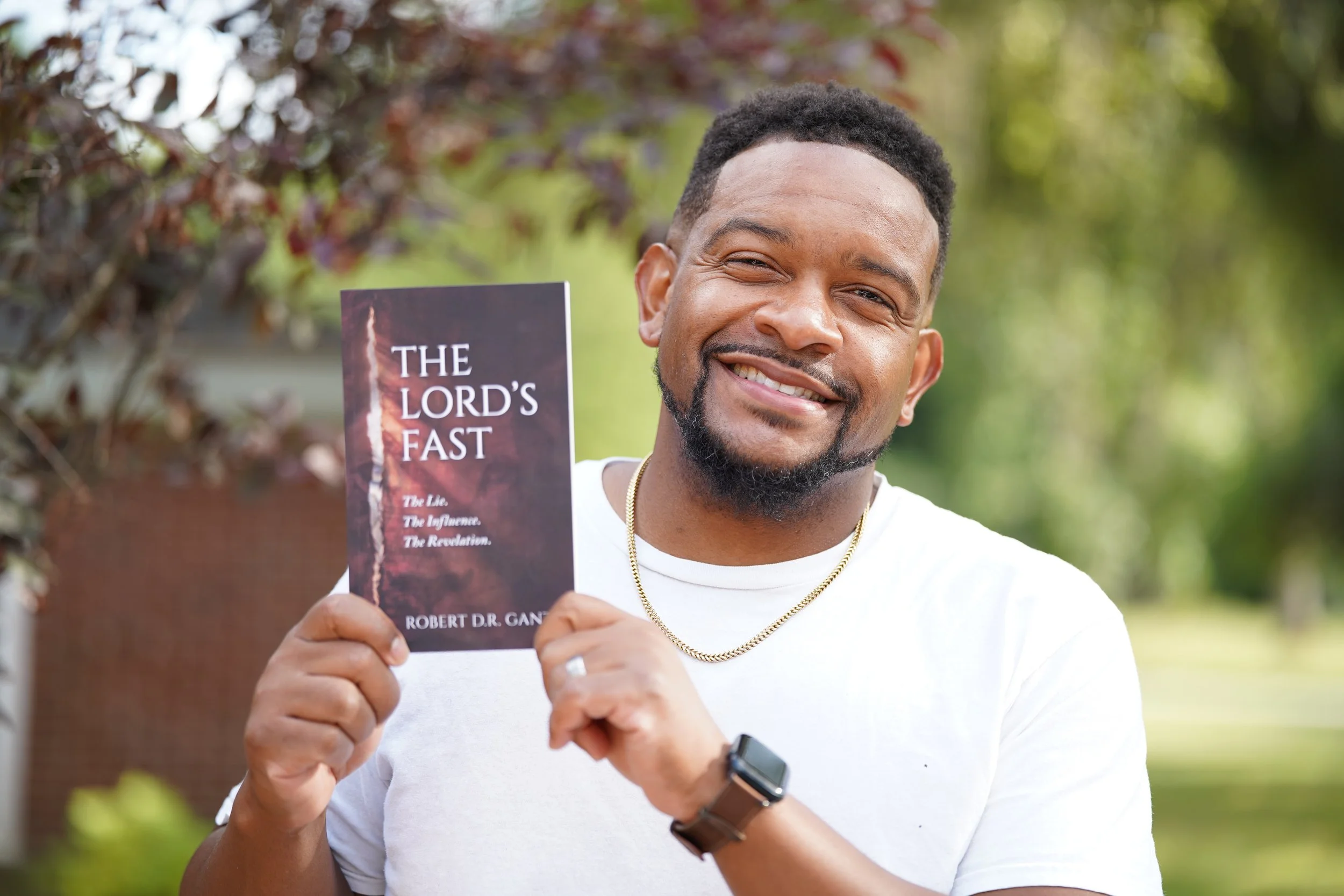 Smiling man holding a book titled 'The Lord's Fast' outdoors among green trees.