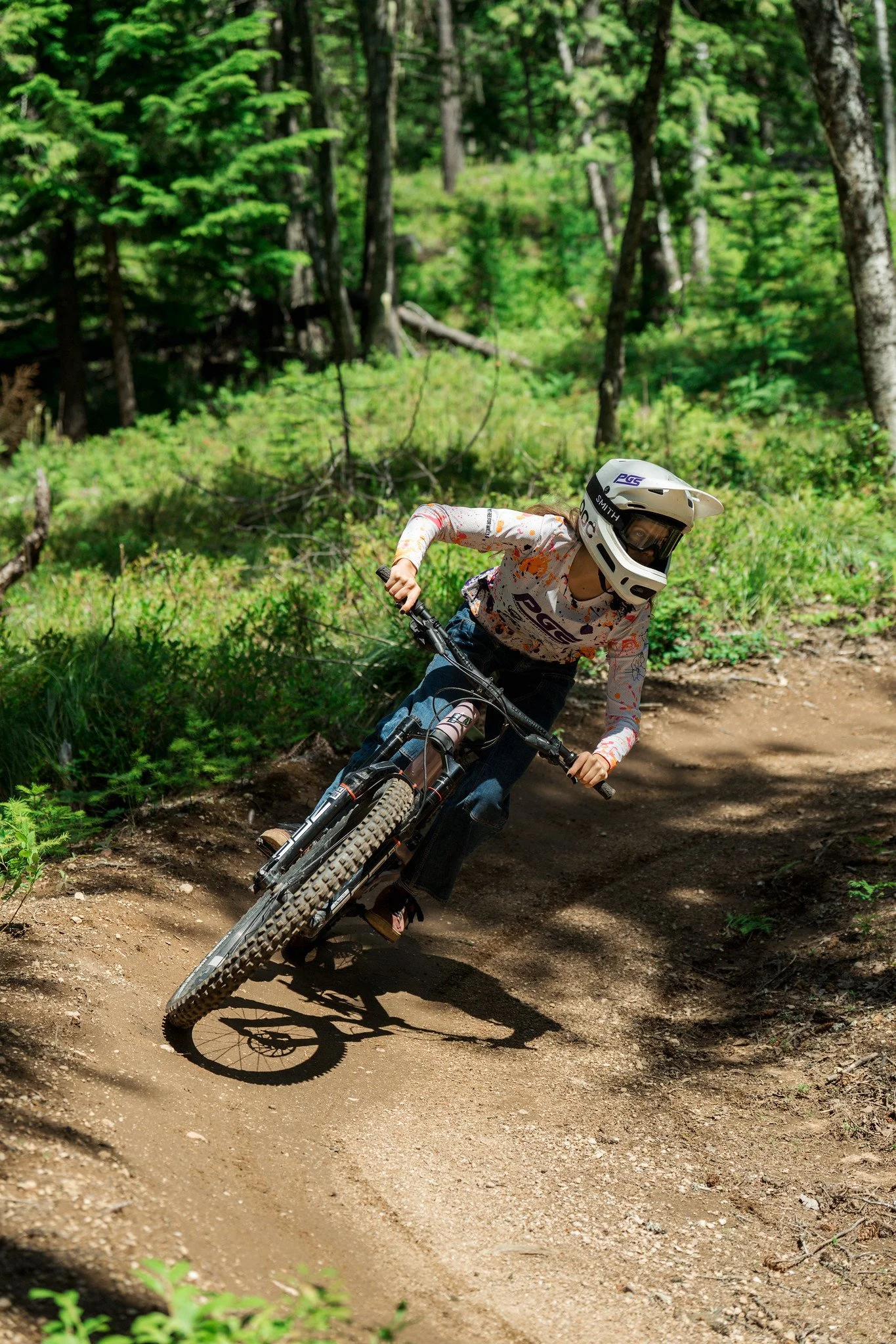 A person wearing a white helmet and a colorful long-sleeve shirt is mountain biking on a dirt trail through a lush green forest.