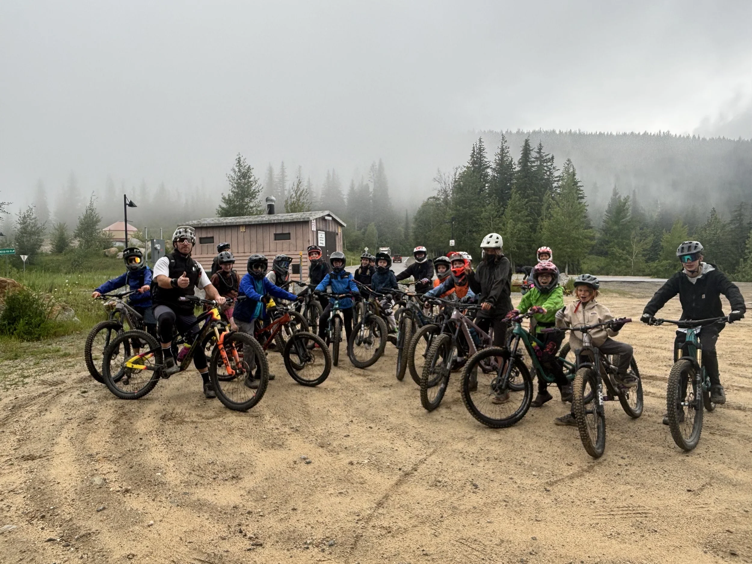 Group of children and adults with bicycles, wearing helmets, gathered outdoors in a scenic area with green trees, a wooden building, and foggy mountains in the background.