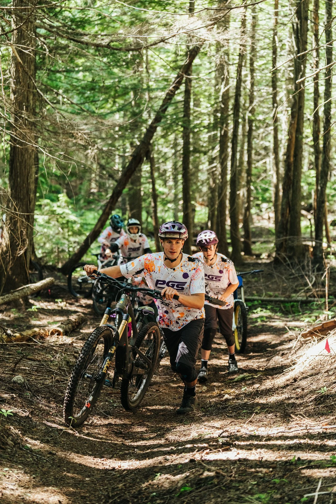 A group of mountain bikers riding on a trail through a dense forest with tall trees and sunlight filtering through the leaves.
