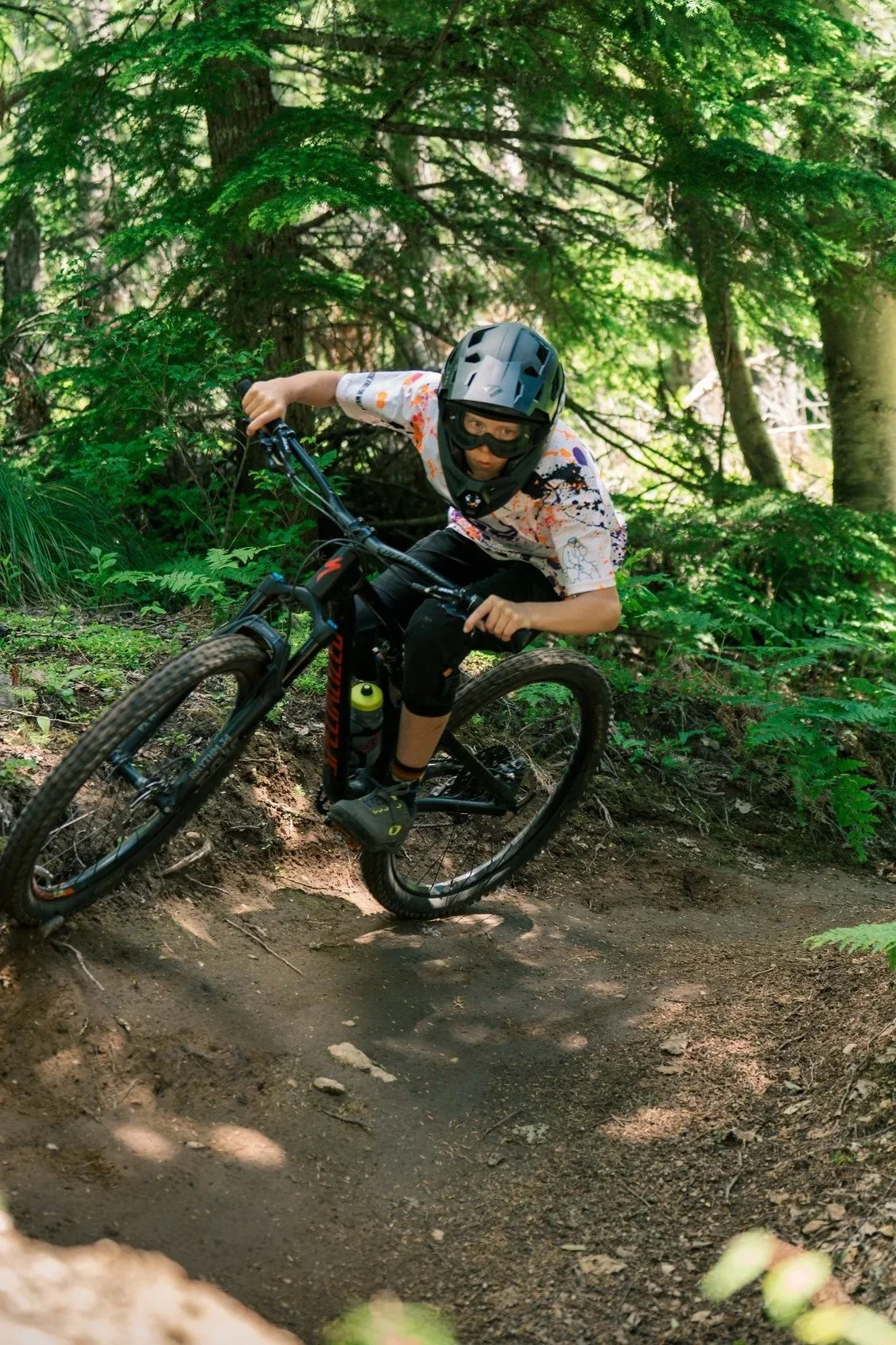 A person mountain biking on a dirt trail through a forest, wearing a helmet, goggles, and a colorful shirt with black shorts.